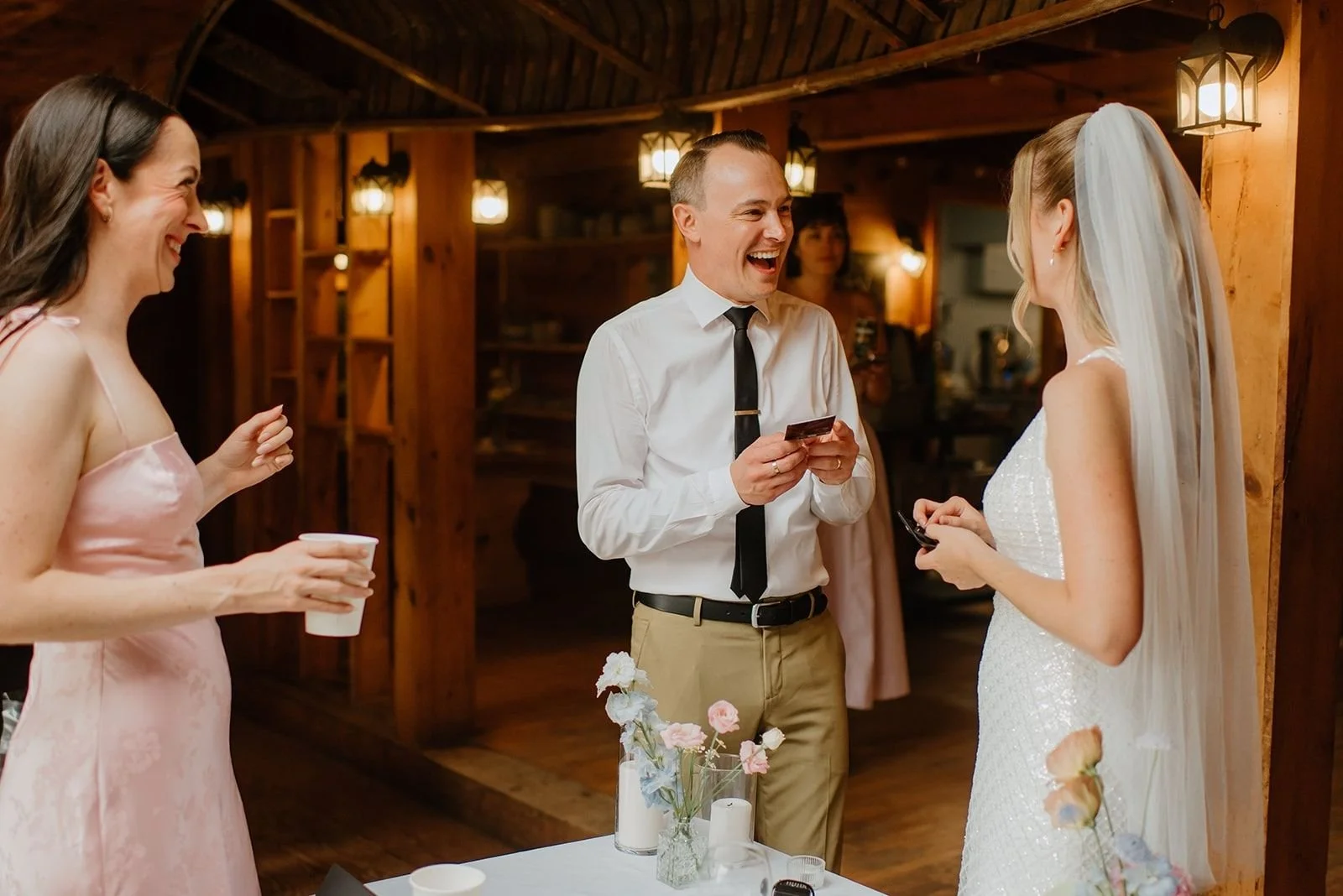 Candid indoor reception moments with Amy laughing heartily while holding a card, surrounded by rustic wood beam architecture and ambient light.