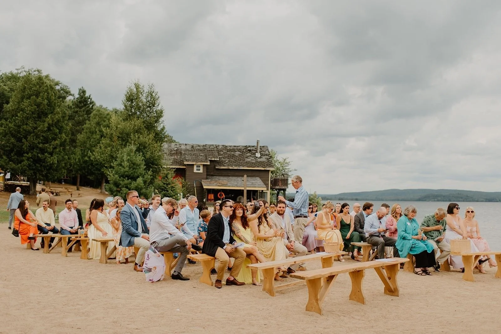 Camp Eden Woods Wedding in Ontario | Amy + Rory – Guests seated on rustic wooden benches at a beach ceremony overlooking the lake in Ontario’s cottage country.