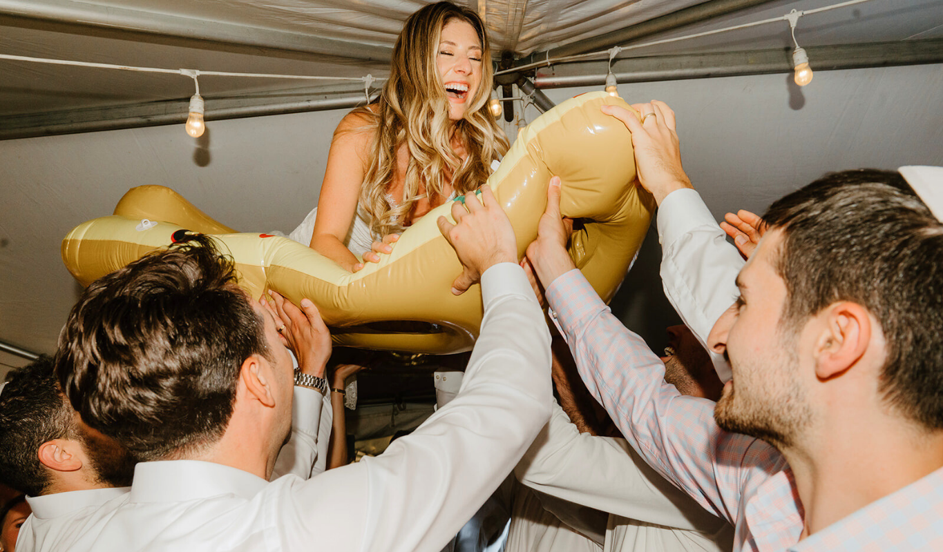Bride crowd surfing on an inflatable float at a wedding reception, surrounded by cheering guests.