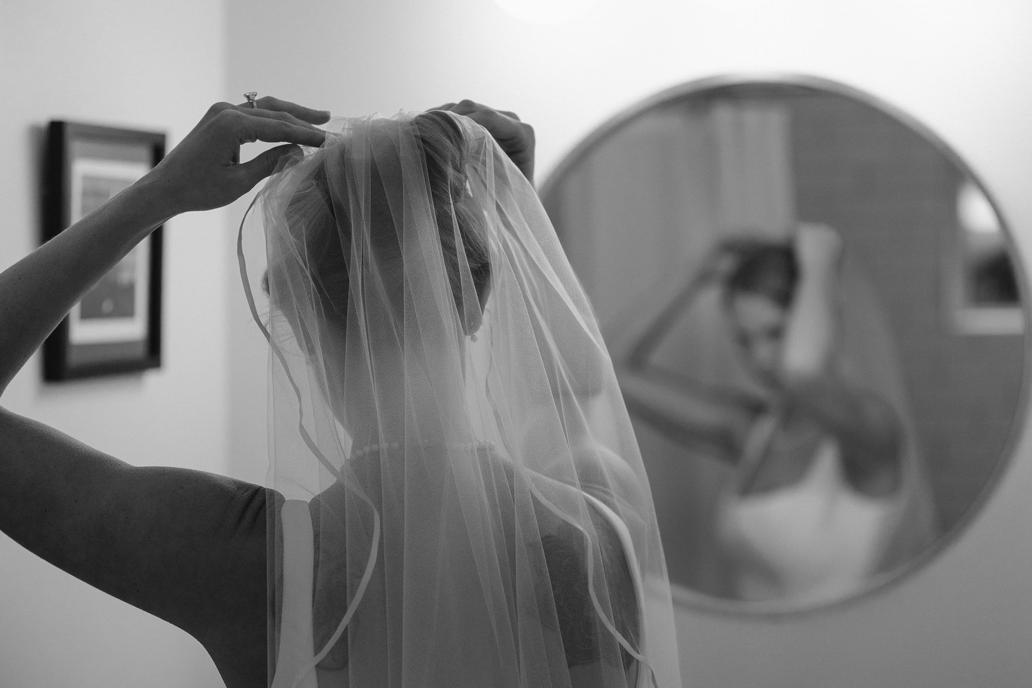 Adding the final touches to her wedding outfit, the bride is seen in front of a round mirror on the wall as she pushes her veil into the top of her updo.