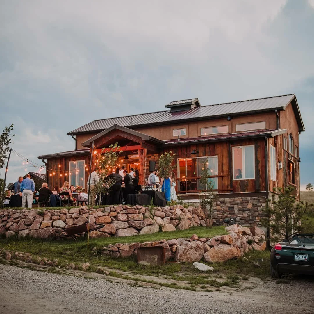 On a summer evening, the 1 C Barn sits stop a stone founation as guests gather in the outdoor reception space on the outside of the building that is decorated with glowing string lights.