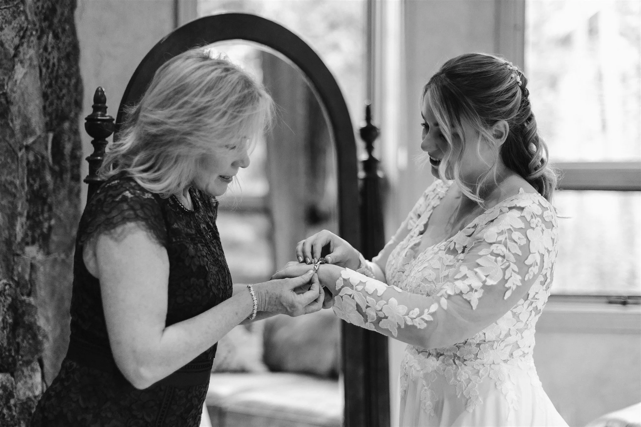 The bride's mother helps her with the final touches as she adds a bracelet to her daughter's wrist . The mother of the bride is wearing a dark lacey dress to compliment the lace and floral theme of the wedding.