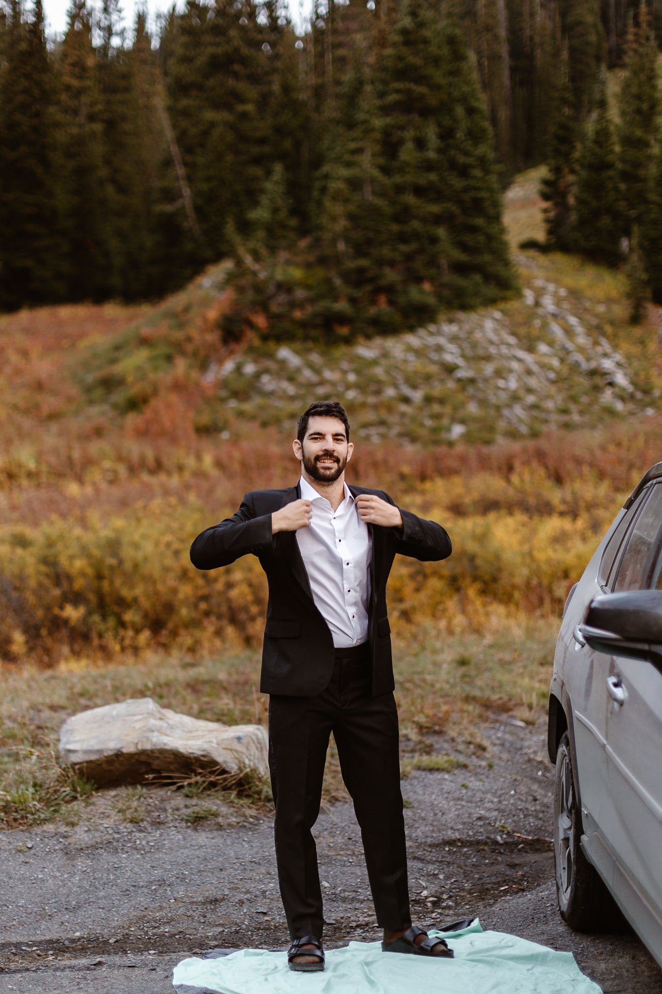 During sunrise portraits at an alpine lake, the groom adjusts his black suit jacket while standing on a green blanket next to their car.
