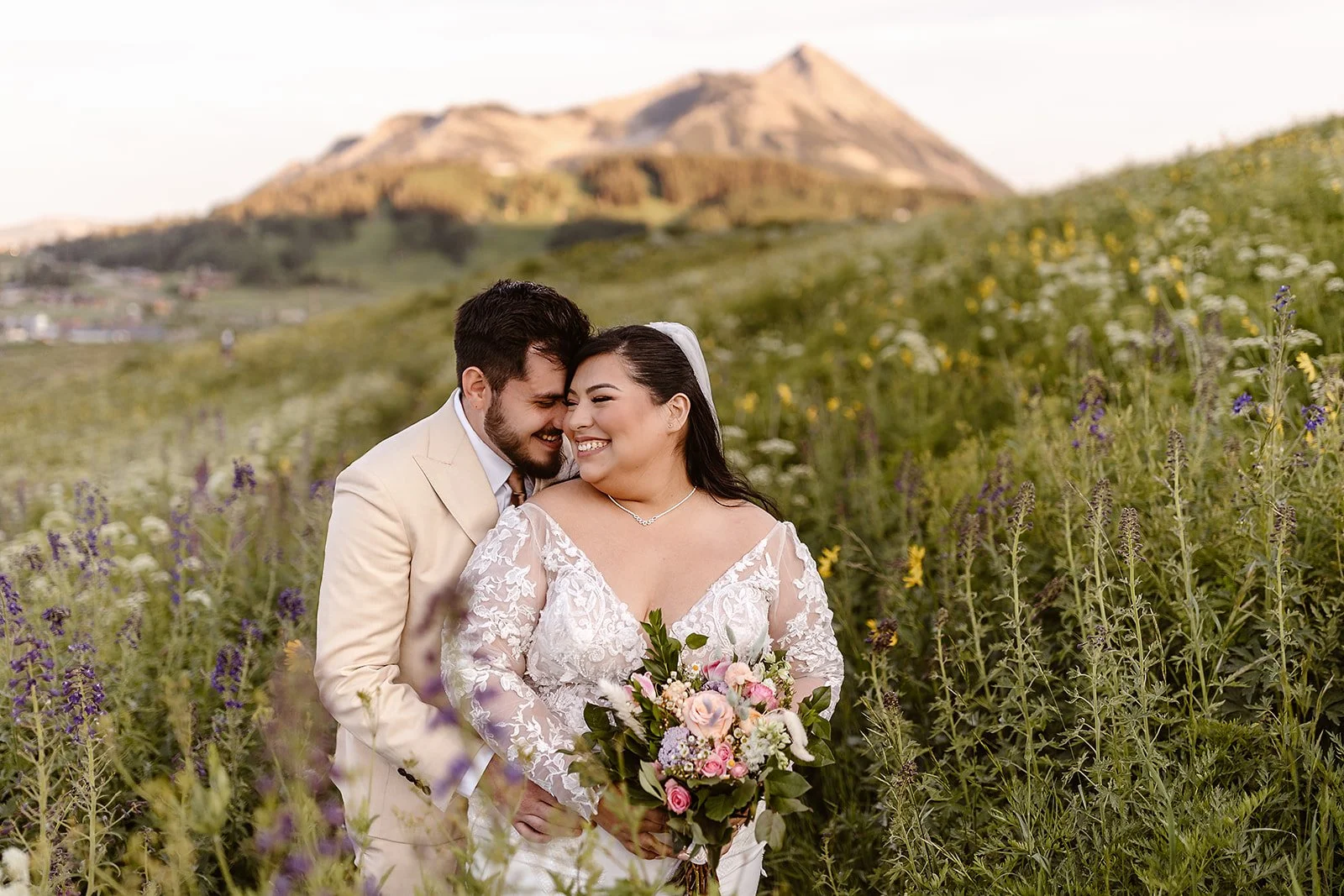 During golden hour, this plus-sized bride and groom are all smiles while surrounded by purple, white, and yellow wildflowers on a warm evening in Crested Butte in July. The groom is holding her from behind, as she smiles widely towards him.