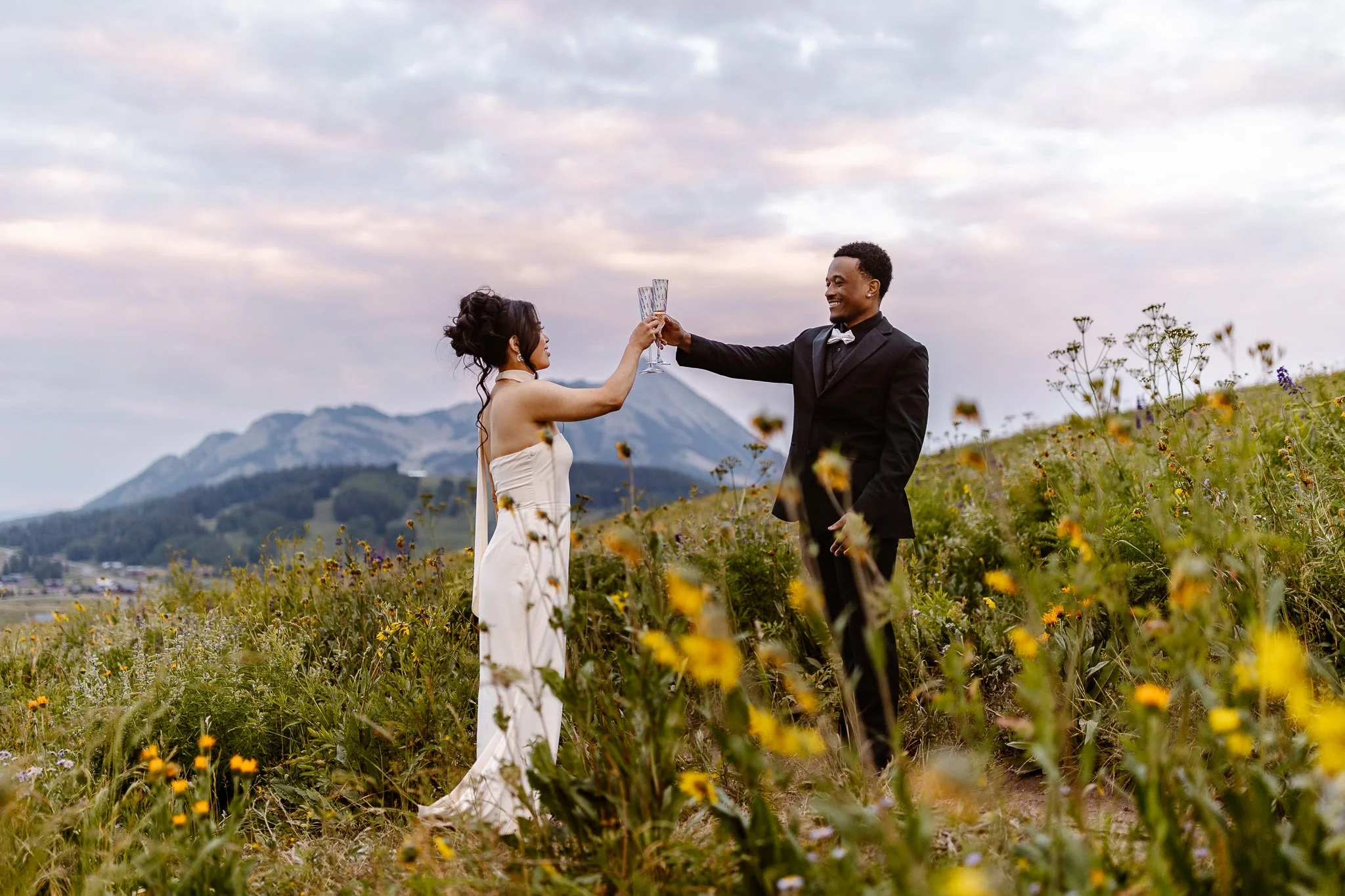 During a colorful sunset amongst the wildflowers in Crested Butte,  the asian bride, in a white sheath dress, and black groom, in a all black suit and white bowtie, hold their champagne glasses together in the air during a celebratory toast.
