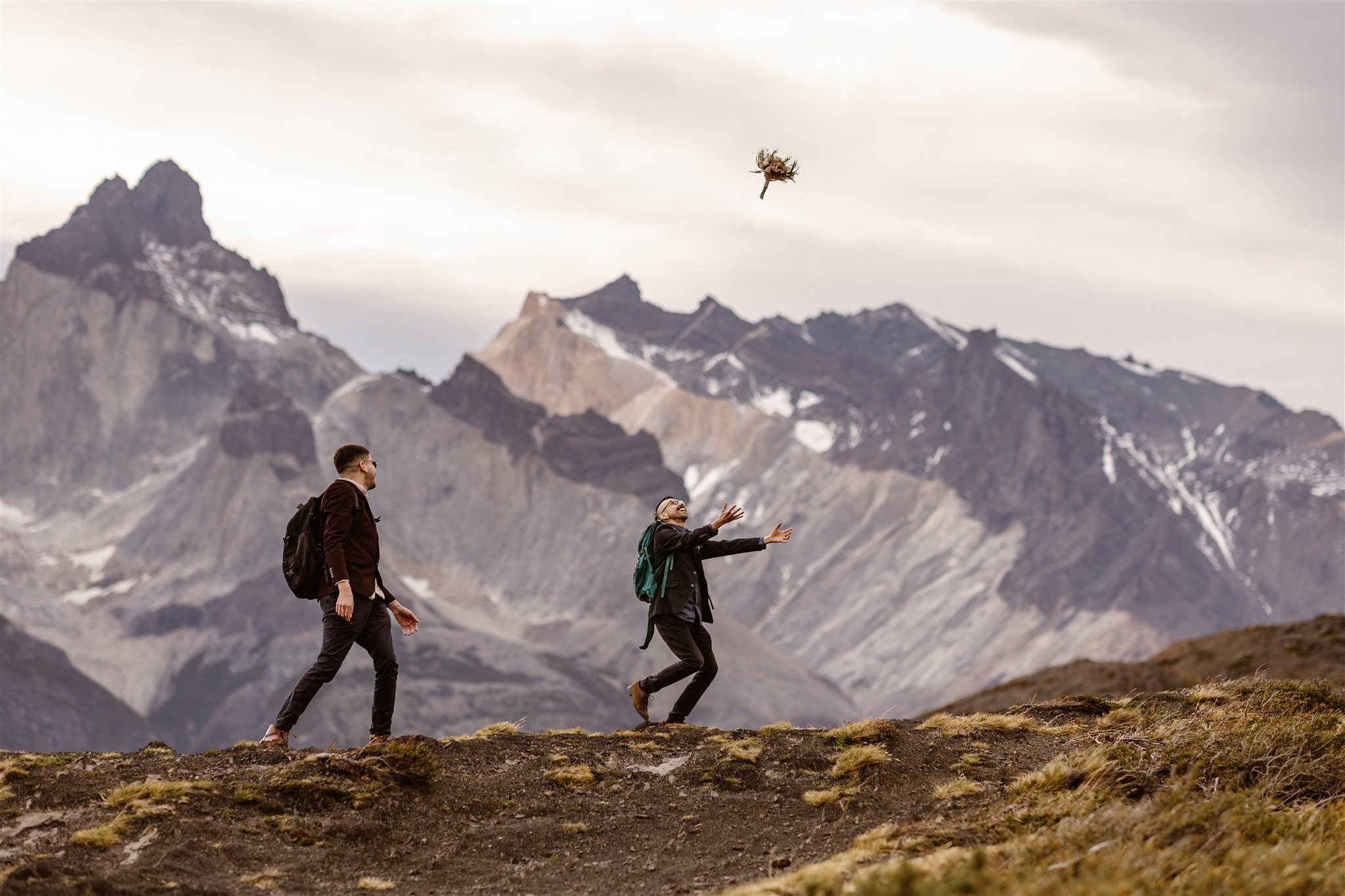 During their hike through Torres del Paine in Patagonia, two grooms enjoy their time together. The one in front is tossing his bouquet into the air while the other watches joyfully.