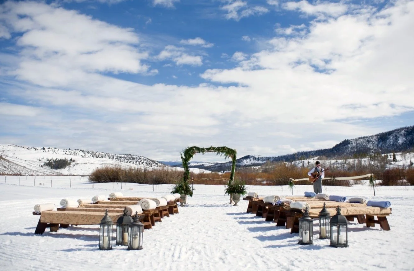 During a snowy winter wedding, there's a ceremony set up in the open lawn of C Lazy U ranch, with wooden benches and blankets set up to create an aisle, with lanterns on the ground that greet guests, and an arch with seasonal greenery at the altar