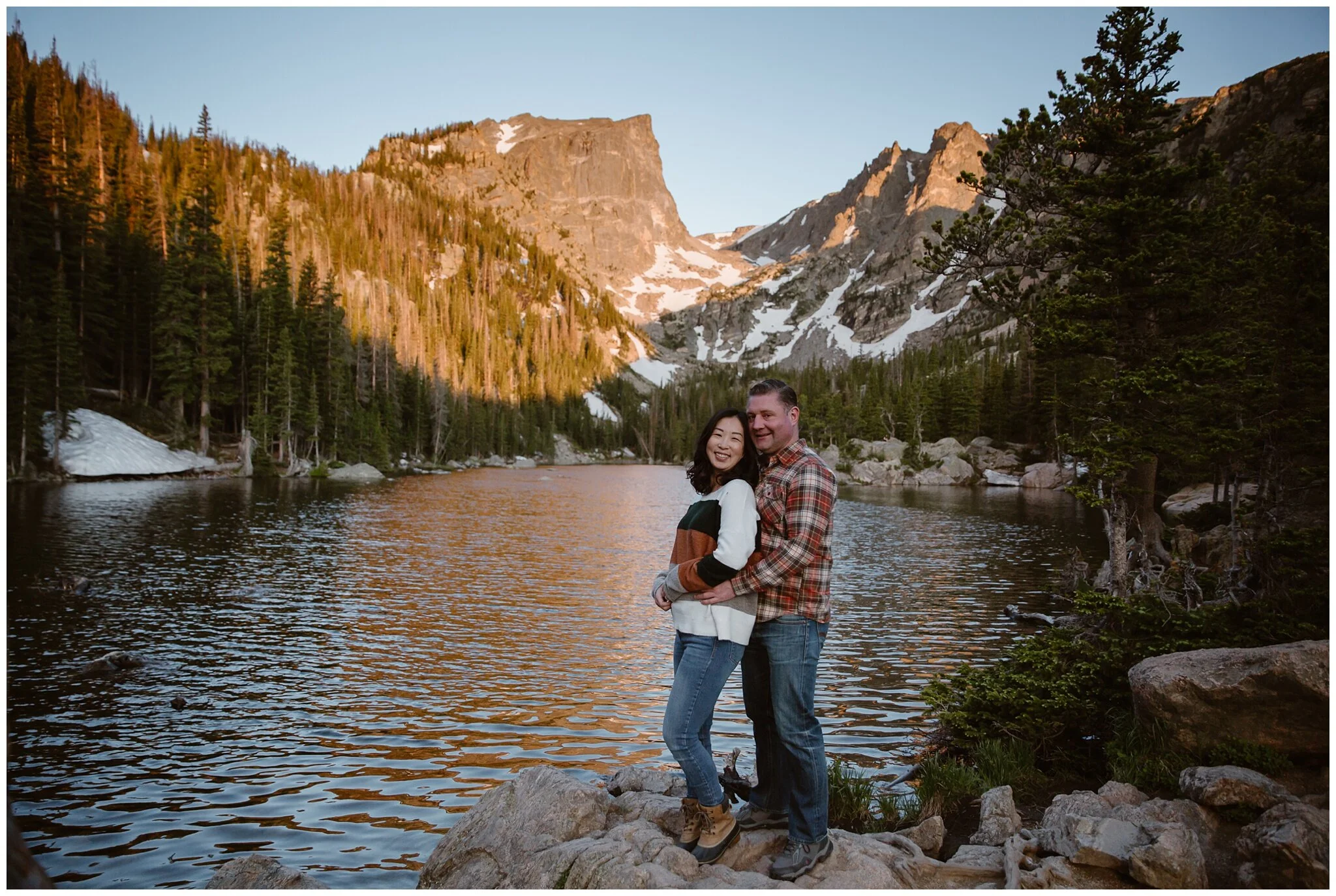 An engagement session in Estes Park, where the couple are dressed in cozy sweaters, jeans, and hiking boots, as they stop on rocks that line the edge of the lake, as the sun sets on the mountains behind them.