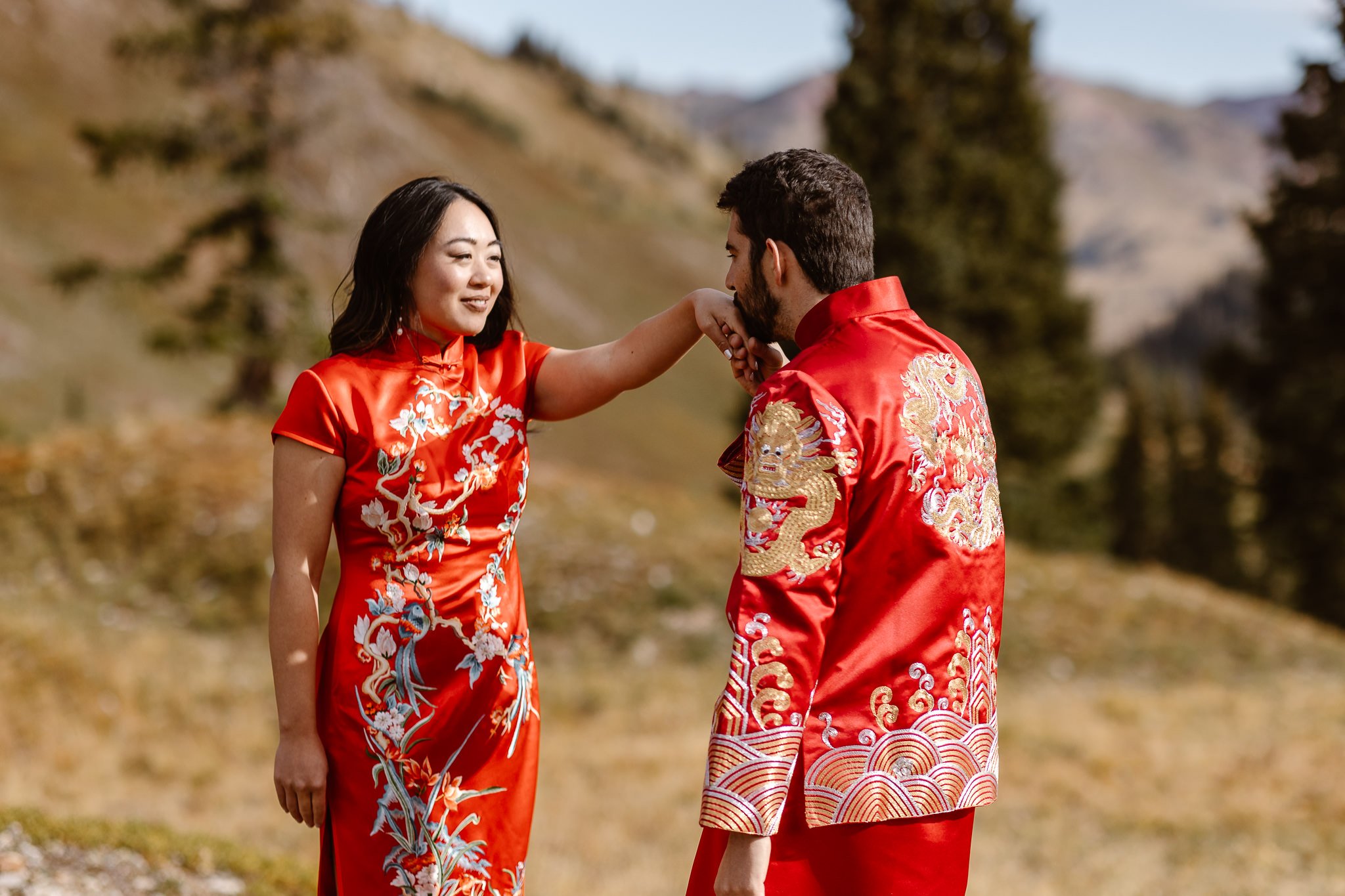 In their traditional chinese outfits, the groom holds his bride's hand and kisses it as she smiles sweetly at him. Their red outfits have beautifully embroidered flowers, birds, dragons, and other intricate details.