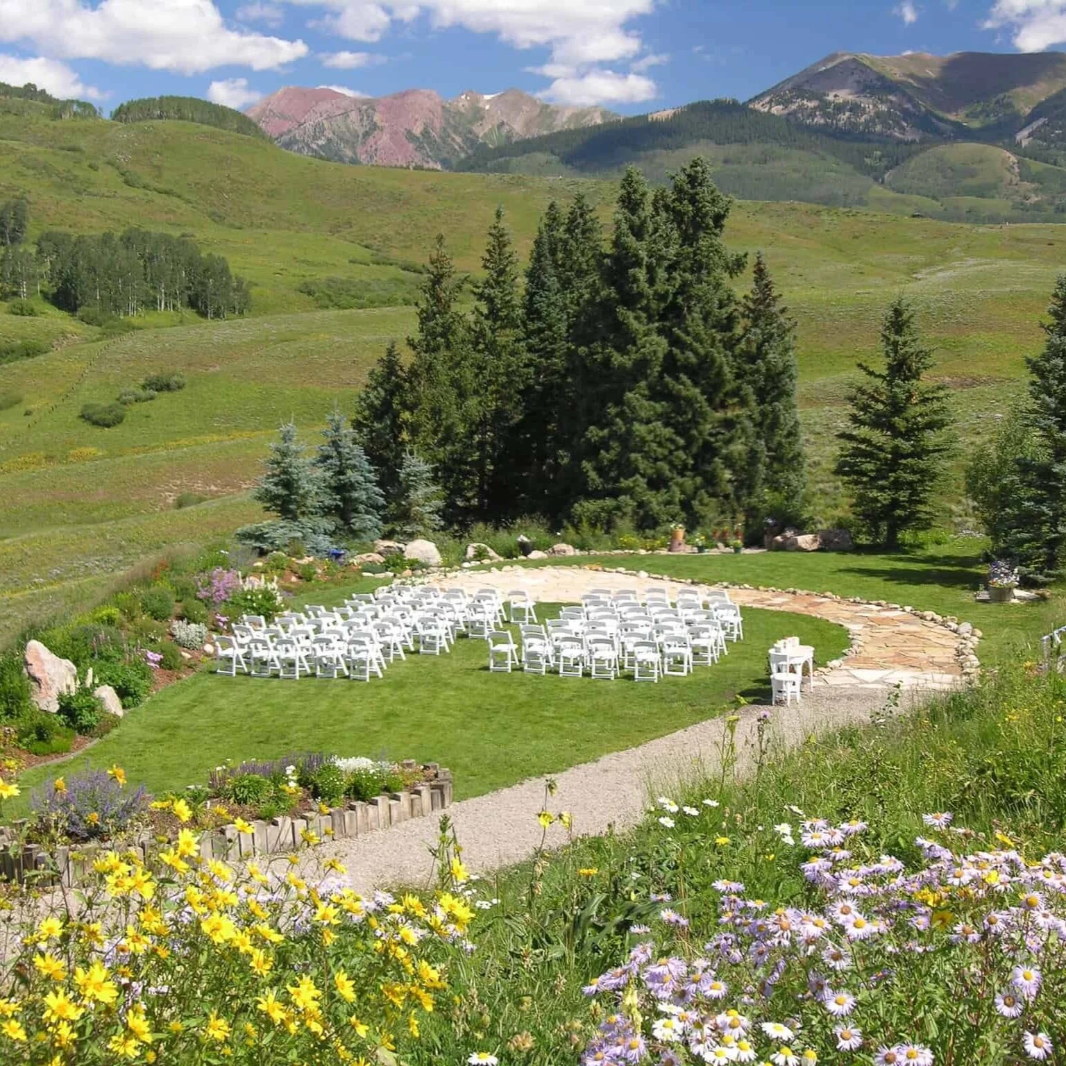 The small gravel path that leads to a curving stone path that serves as an aisle for weddings at this outdoor ceremony space. There are white chairs arranged on the grass area that look upon the stone path and rusty red mountains of Crested Butte.