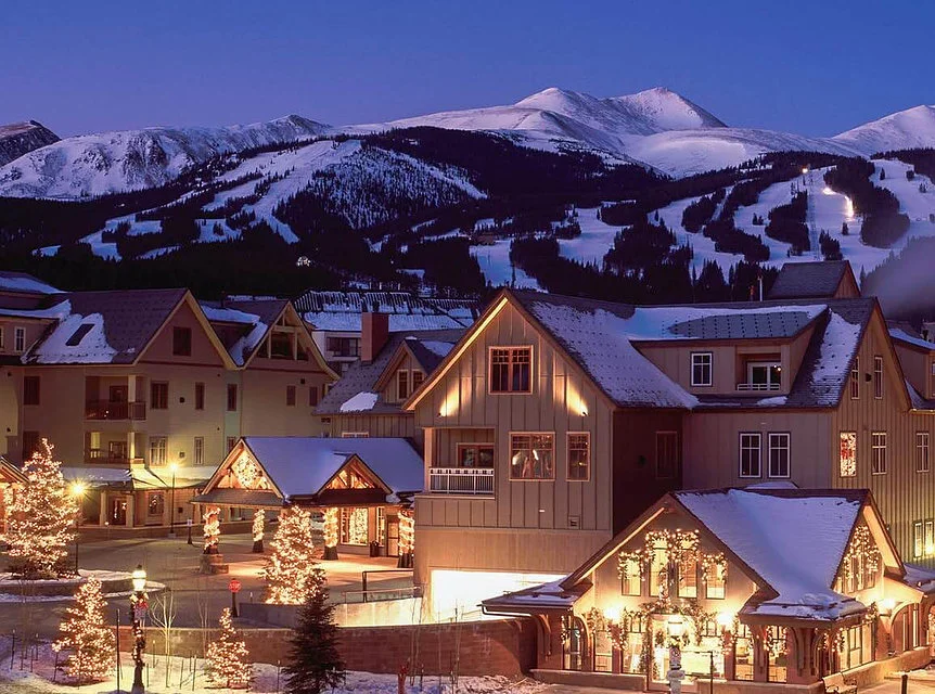 A night view of the exterior of Main St Station in Breckenridge during the holidays, as Christmas lights illuminate the trees and wrap around the poles and windows, showing the snow filled mountains in the near background.