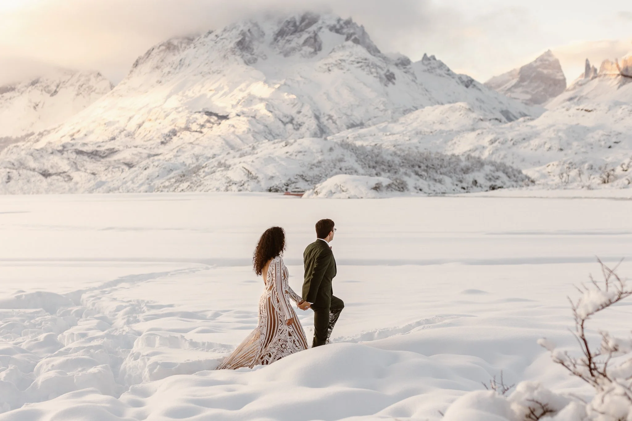The bride holding the grooms hand as he leads her through the snow on the morning of their winter micro wedding in Patagonia. Her dress is a unique pattern and he dons a green suit that pops against the white of the snow covered ground before them.