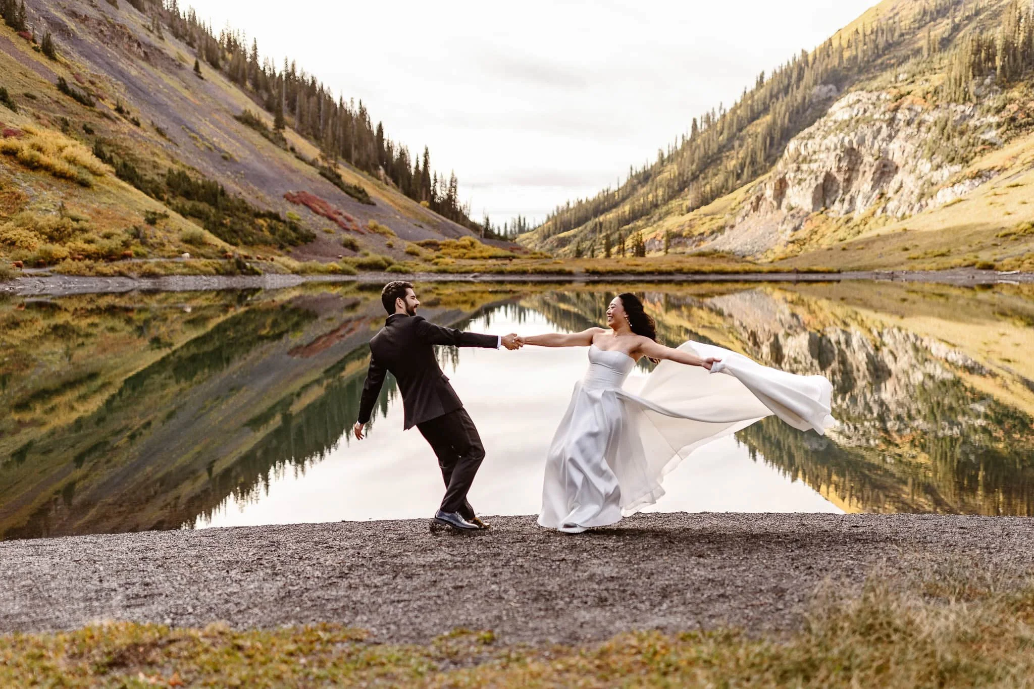 The bride and groom hold hands while spinning in a circle, her dress flying in the wind behind her, both smiling joyously during this beautiful sunrise moment.