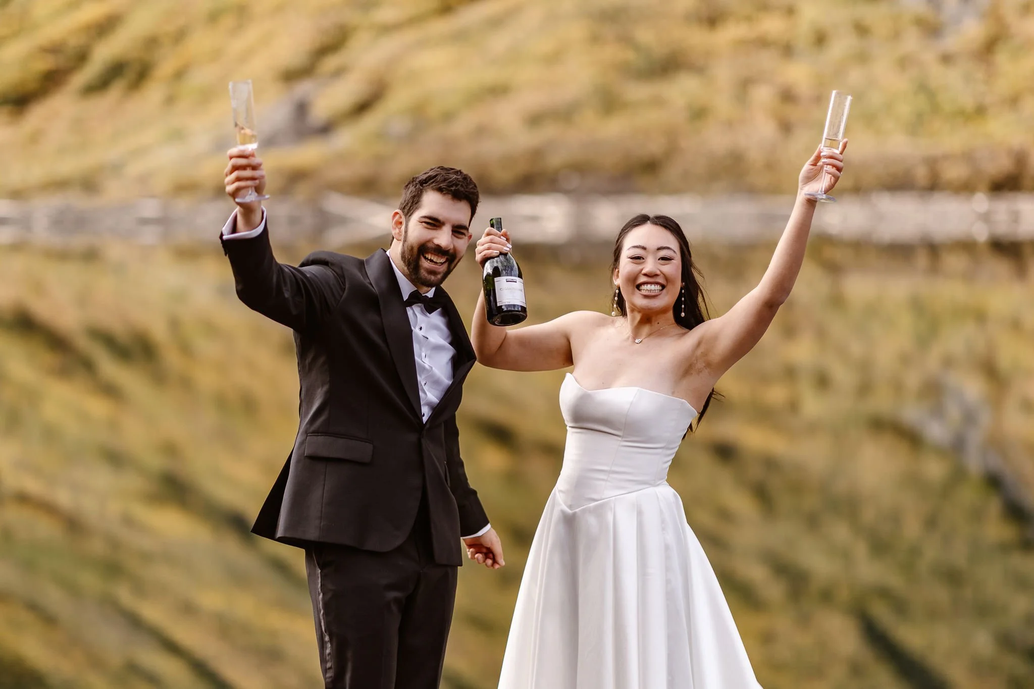 Colorado elopement photographer Andrea Enger captures a groom and bride smiling  with their hands in the air as they each hold a champagne glass and the bride holds the bottle in the air.