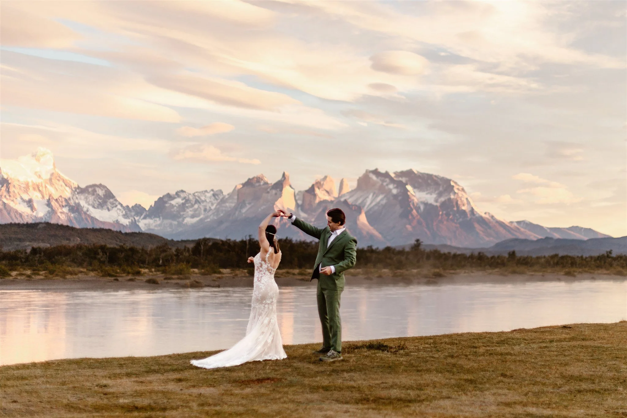 An eloping couple dancing on the edge of the water during a pink sunrise in Torres del Paine, Patagonia. The groom is twirling the bride as they dance in celebration.