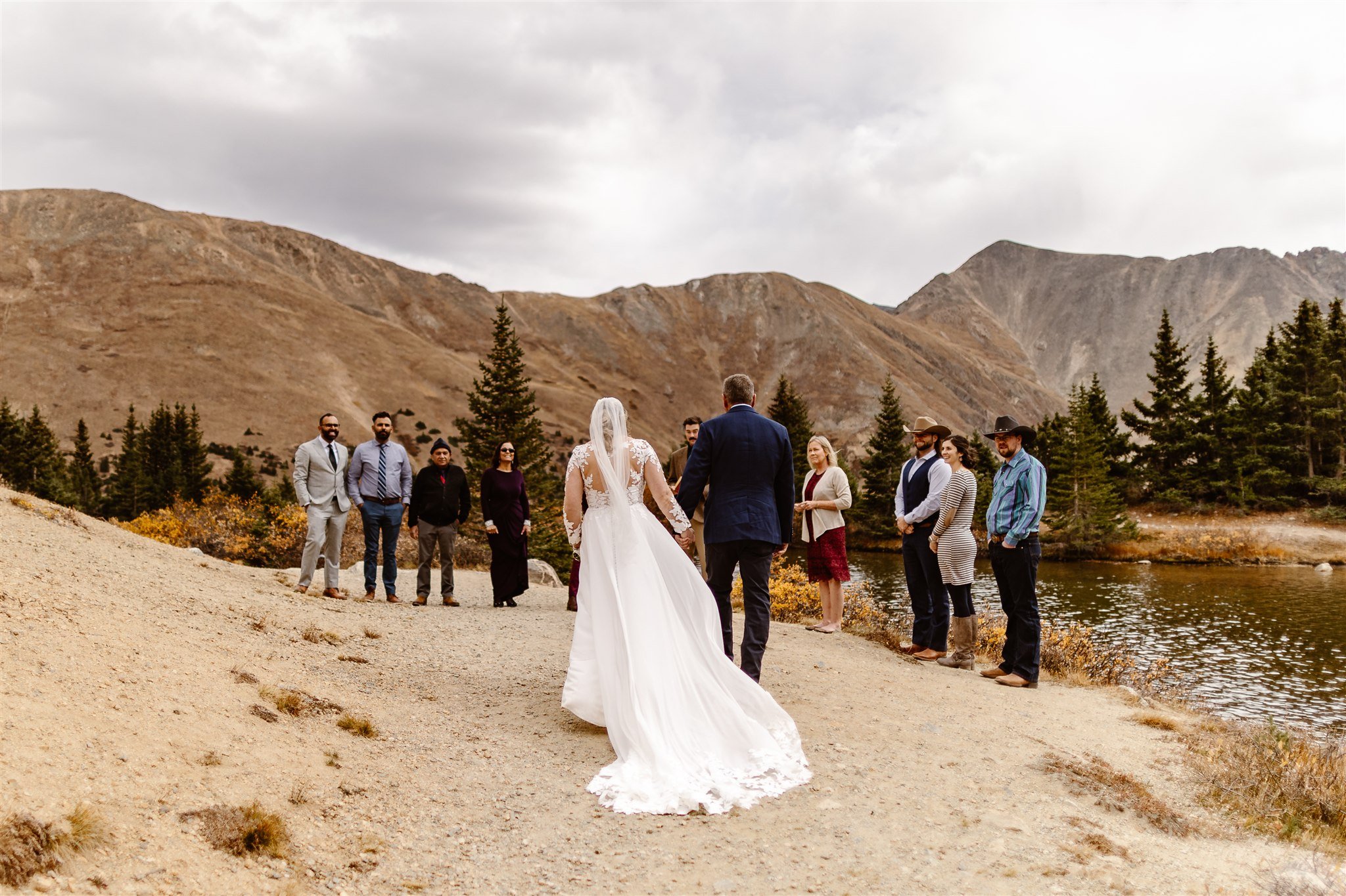With the brown and grey mountains of Breckenridge and calm lake as their backdrop, the bride walks along the shore of the lake towards their wedding guests while holding her dads hand. The guests are standing in front of the mountains as they wait.