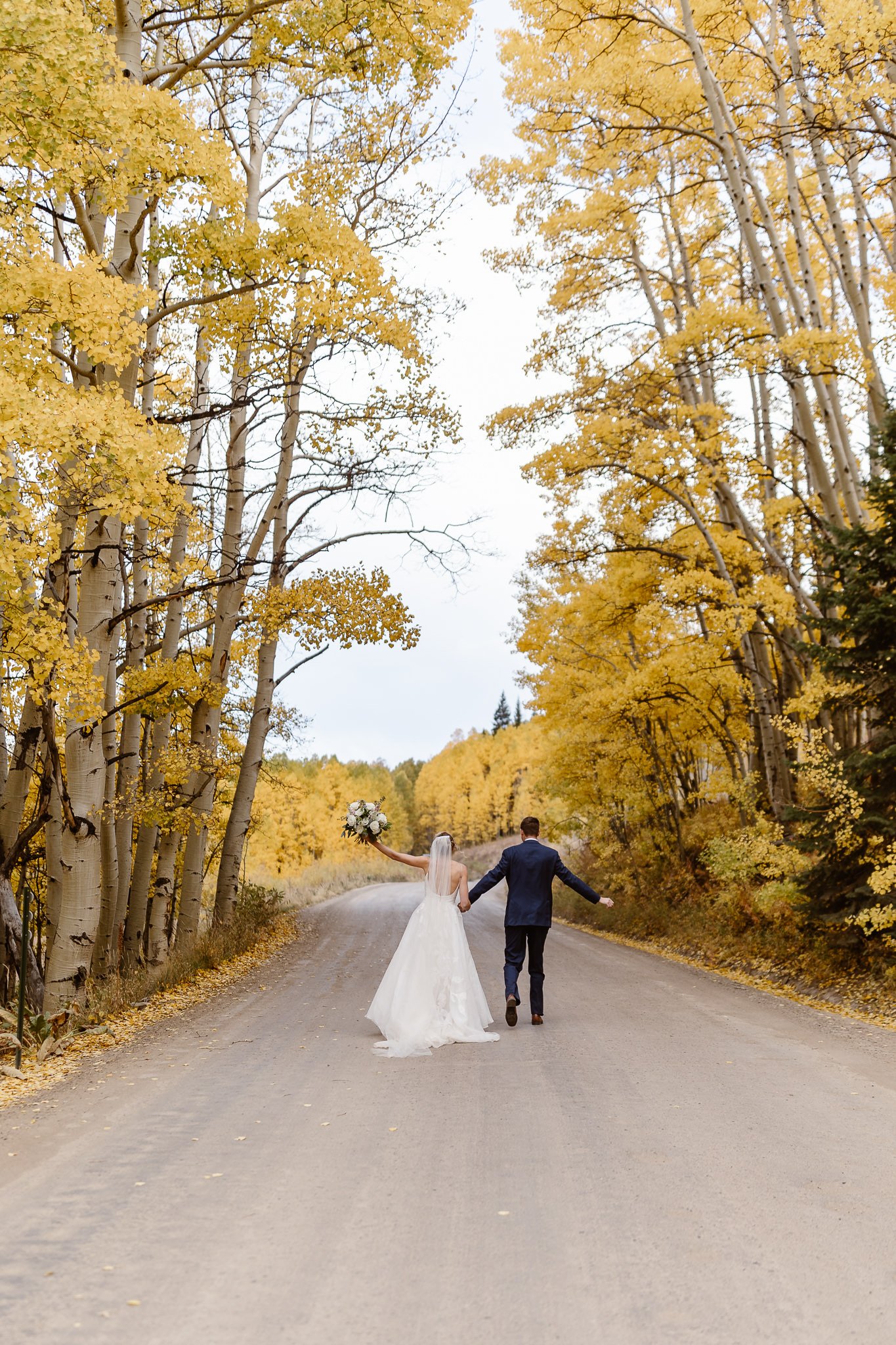During their September micro wedding in Crested Butte, newlyweds hold hands while running down a paved road that is surrounded by bright yellow aspen trees.
