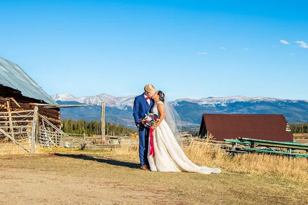On a sunny afternoon at Snow Mountain Ranch, a bride and groom kiss outside of the barn buildings on one of the wedding spaces available on the property. The sky is clear and you can see the details of the mountains that fill the background.