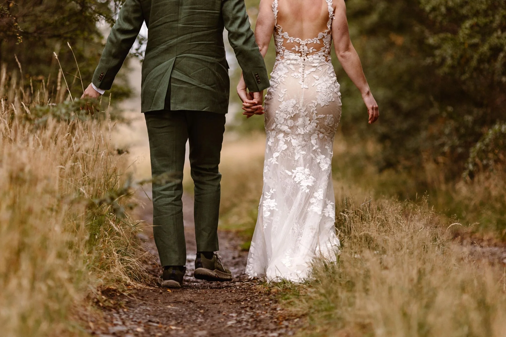 Shot from behind, the bride and groom hold hands as they walk a dirt trail to their ceremony spot by a different part of the water.