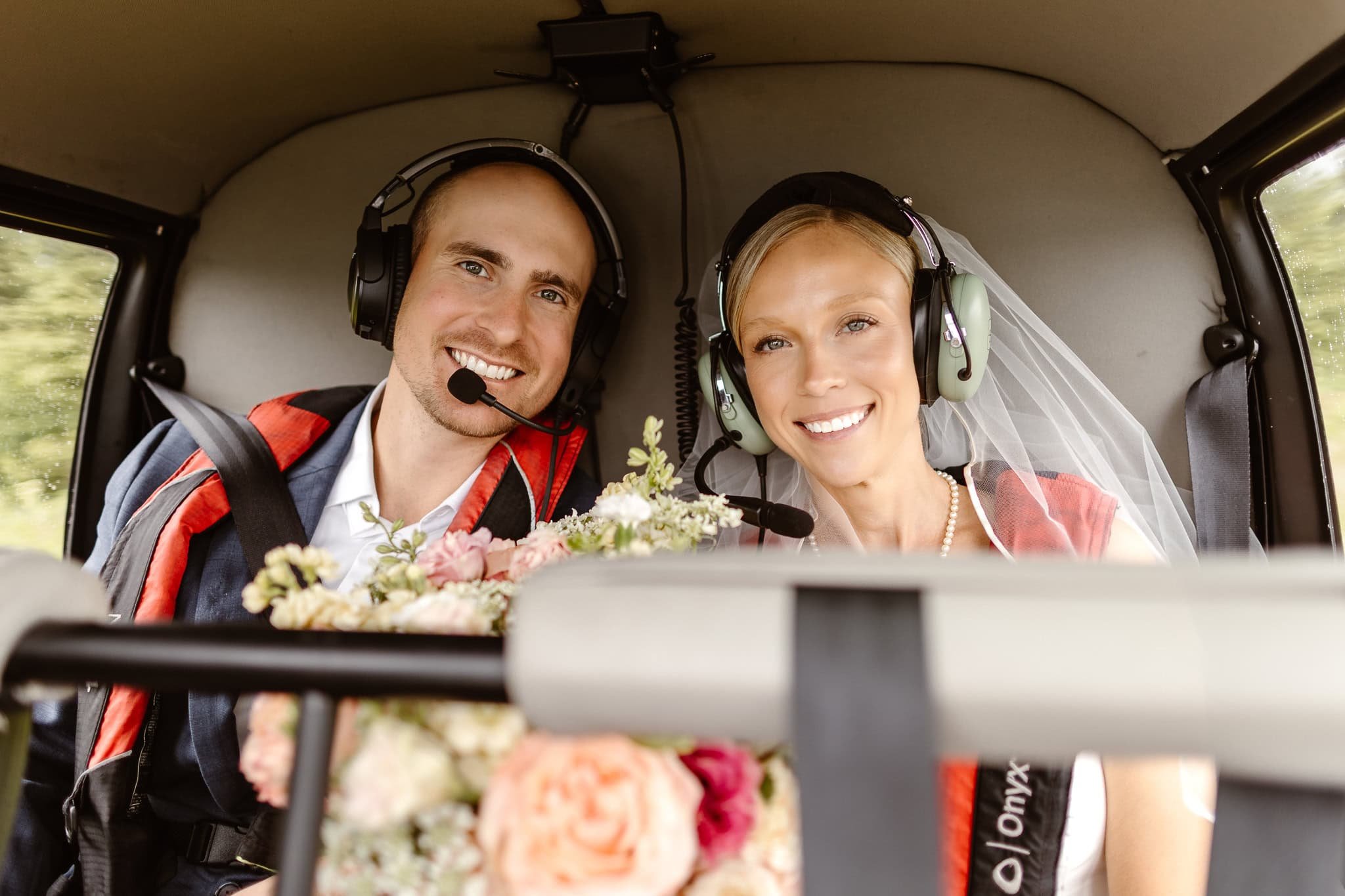 On their way to the glacier, the bride and groom smile joyfully during their helicopter ride. Each wearing a headset and red safety harnesses.