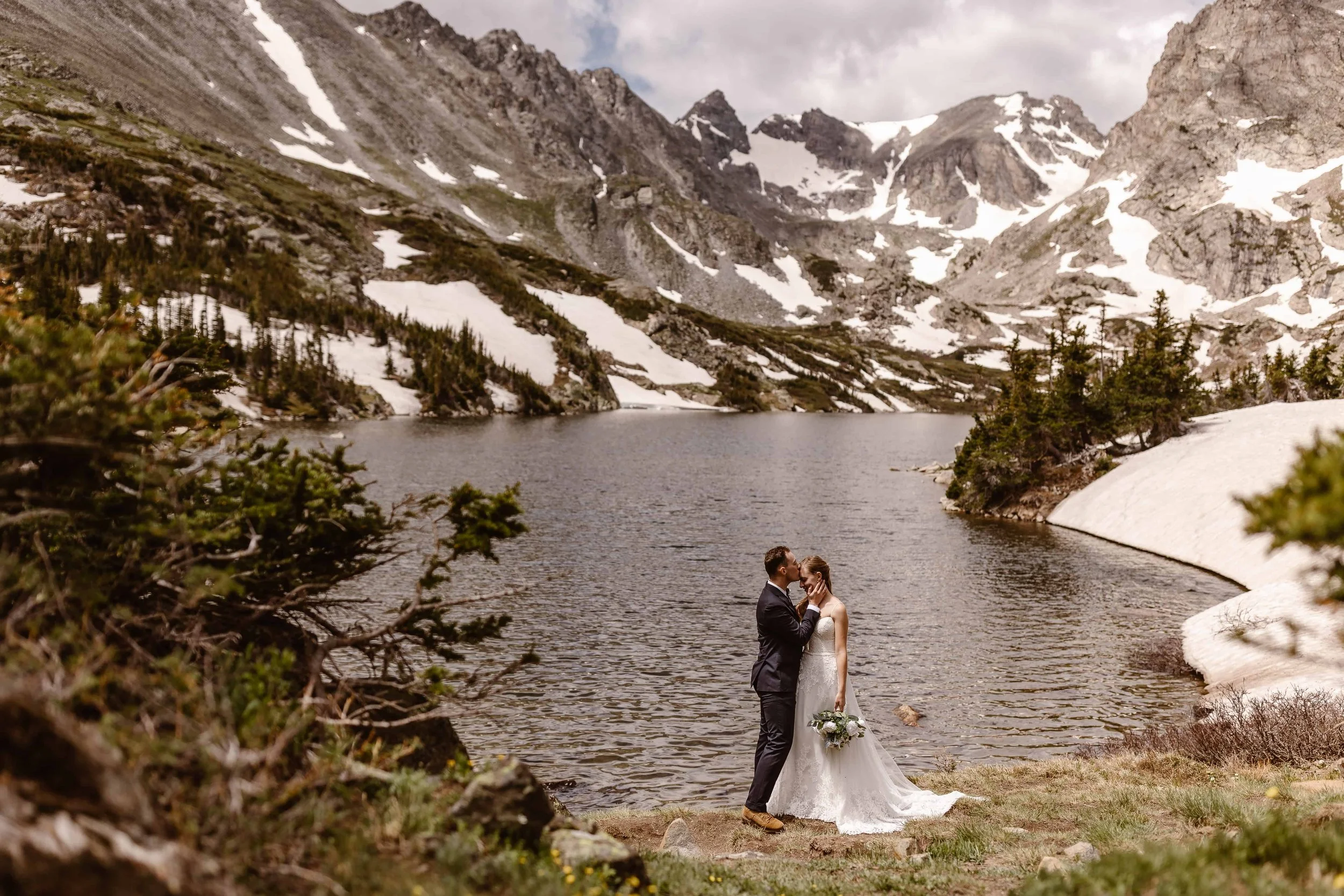 During this afternoon elopement, the bride and groom stand on the very edge of Lake Isabelle, as the groom kisses his bride's forehead and she holds her white and green bouquet in her left hand. There's still snow on the mountains and rocks.