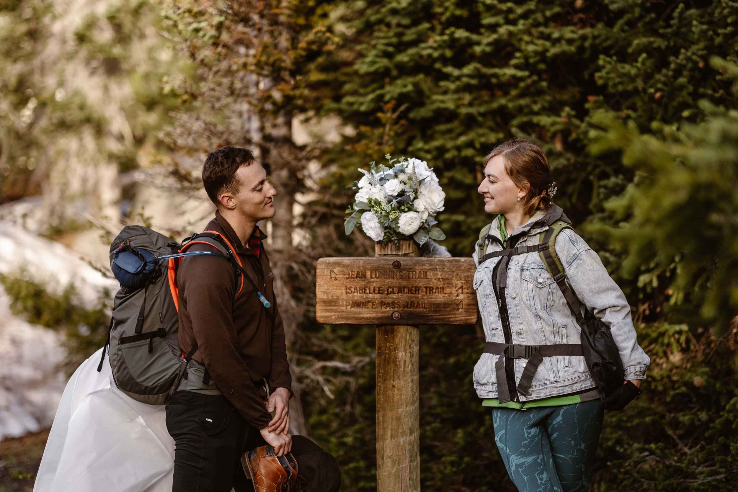 The eloping couple stop to take a photo with the wooden trail sign that leads to Lake Isabelle. The bride on the right and the groom on the left, both looking at each other lovingly.