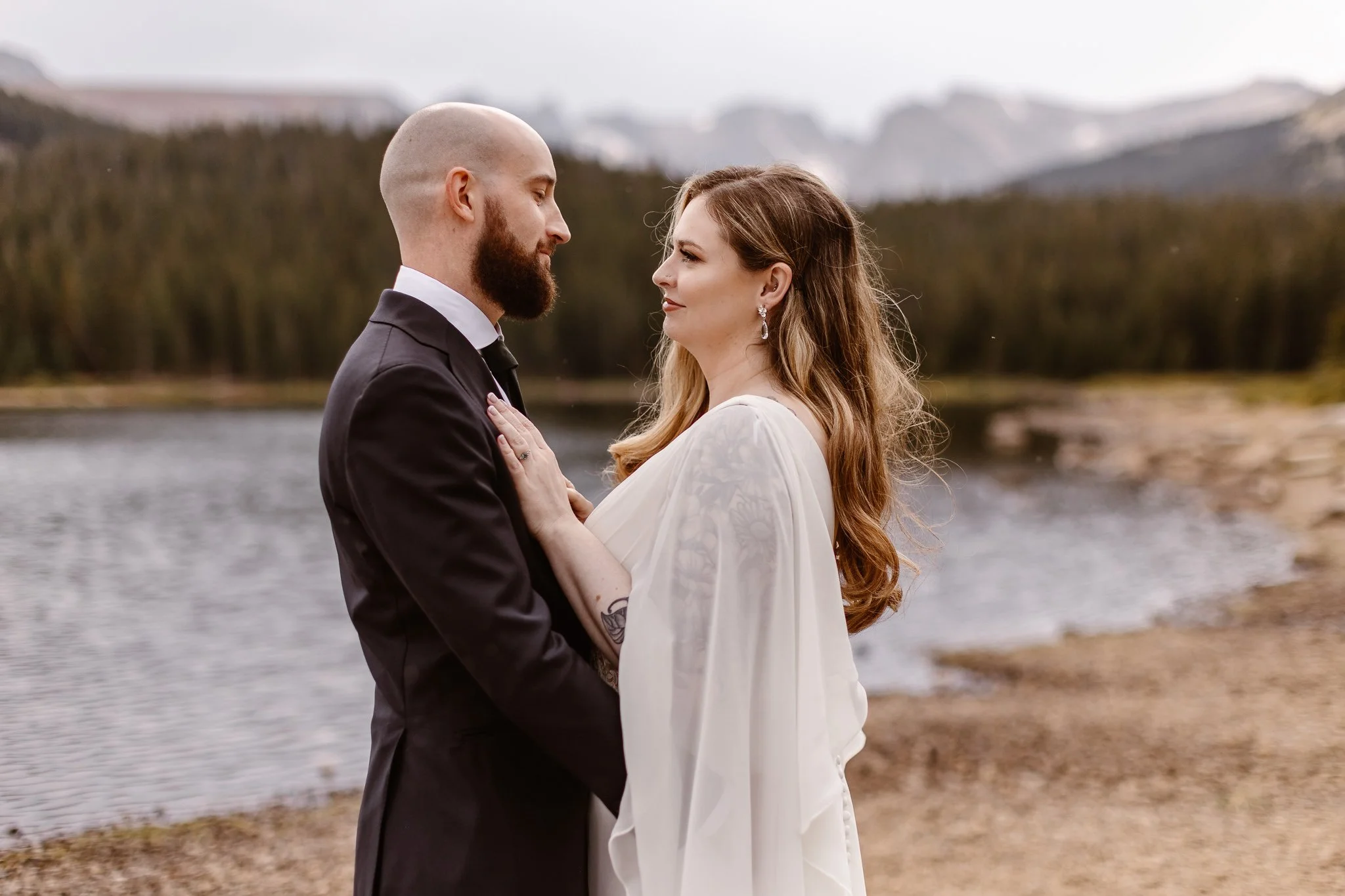 On the rocky lake shore, the bride rests her hands on her groom's chest as they look deeply into each other's eyes.