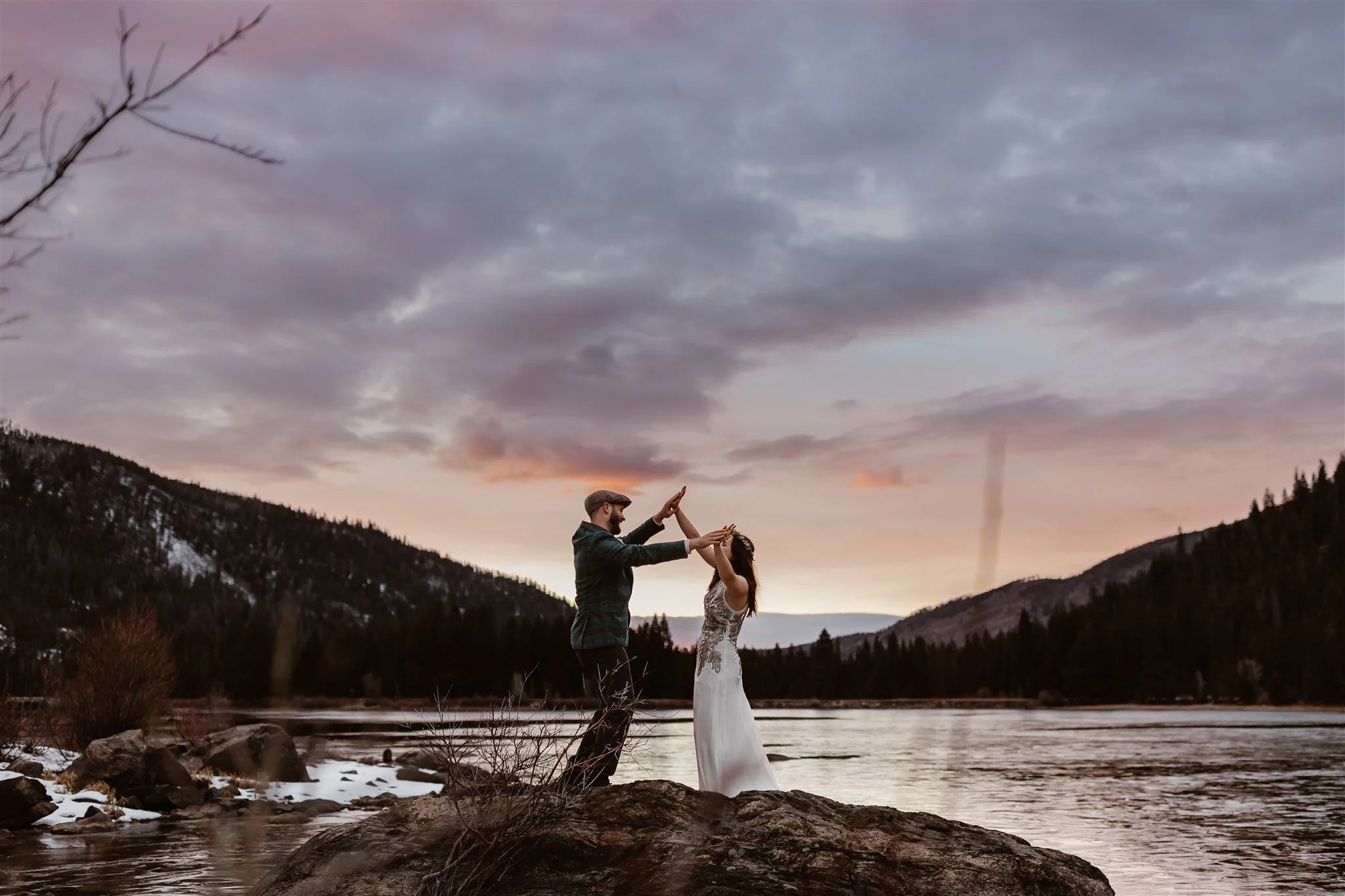 During a sunset first dance at Grand Lake, CO, the newlyweds dance on a rock on the lake with their hands in the air, palms touching. The sky behind them is painted with pastel blue, purple, pink, orange, and yellow as the sun sets.