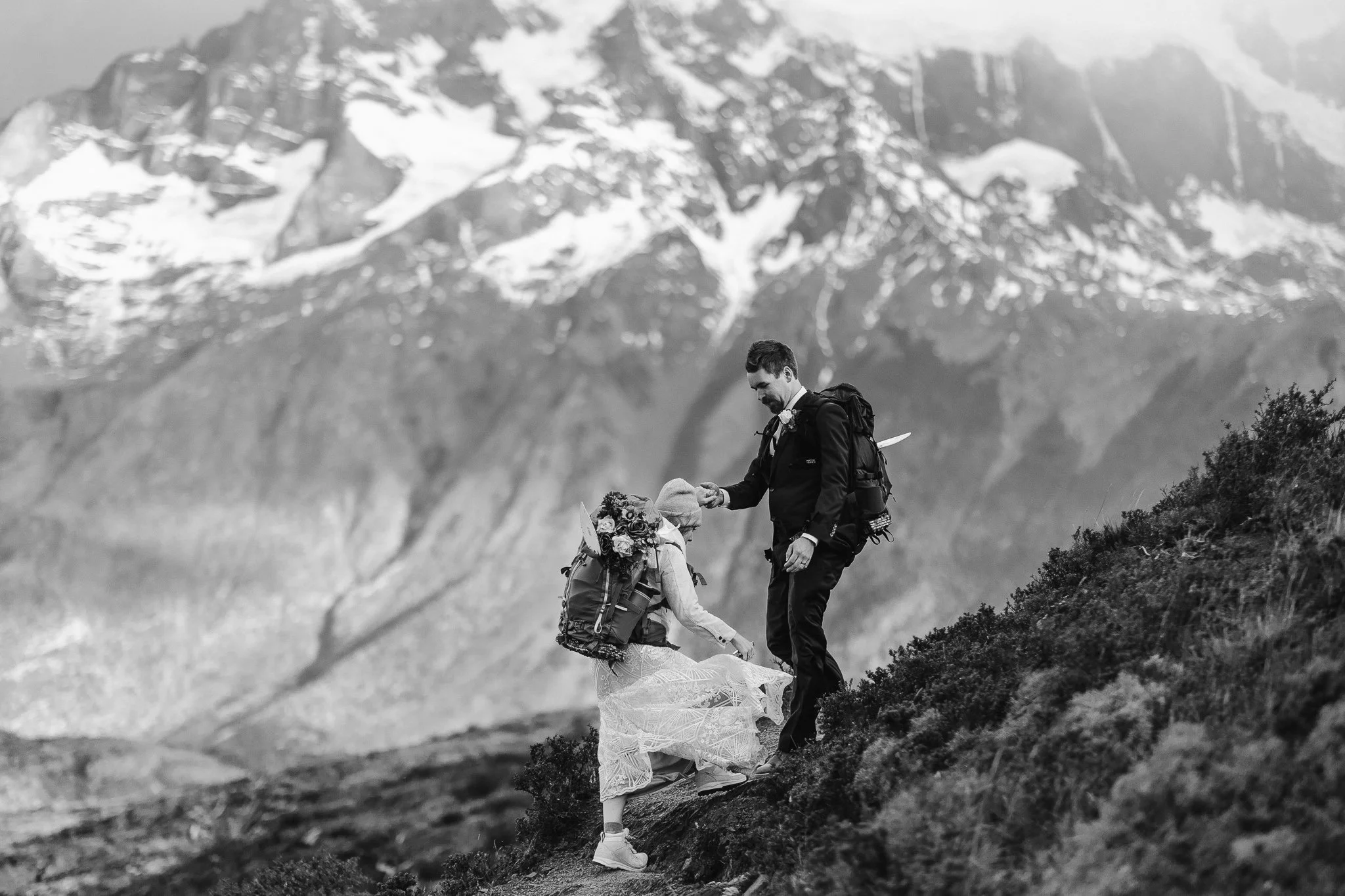 During their hiking elopement through Torres del Paine, the groom helps his bride step up onto the trail and she takes her time making to ascent. The huge mountains fill the background, revealing pockets on snow throughout the peaks.