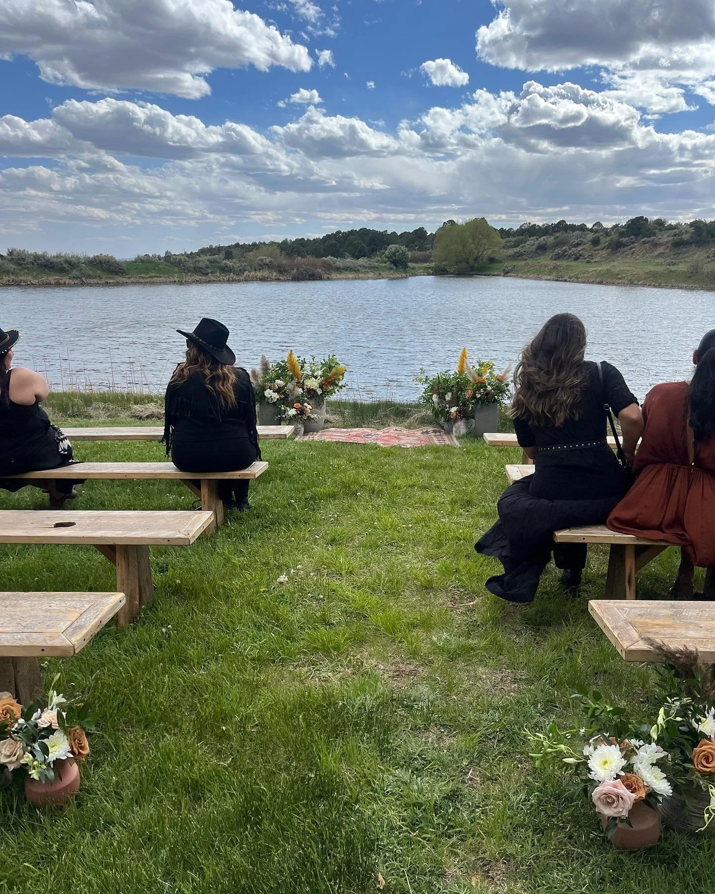 A wedding ceremony at the blue lake of Blue Lake Ranch, with wooden benches set up on either side of the grass aisle and a rug with floral arrangements on the ground at the altar, right next to the lake.