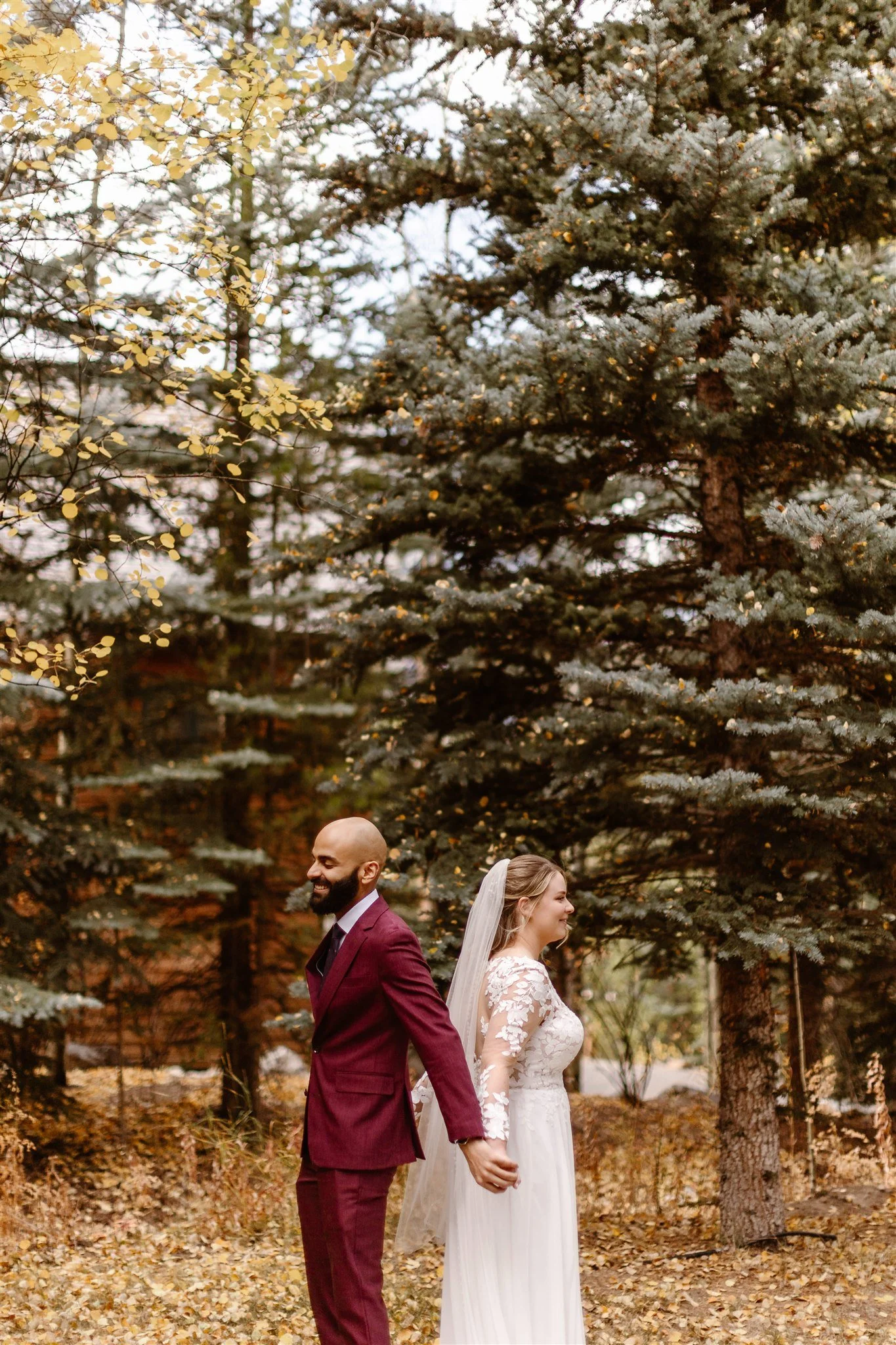 During their first look under an aspen tree, the bride and groom stand back-to-back while holding hands, both smiling in anticipation before seeing each other in their wedding outfits.