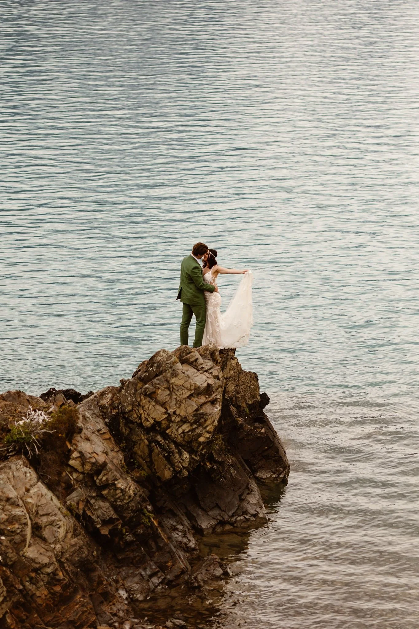 A closer image of the bride and groom standing on the edge of this rocky outstretch on the water as she holds her dress train in the air and he holds her closely from her waist, a romantic moment indeed. The blue of the water ripples around them.