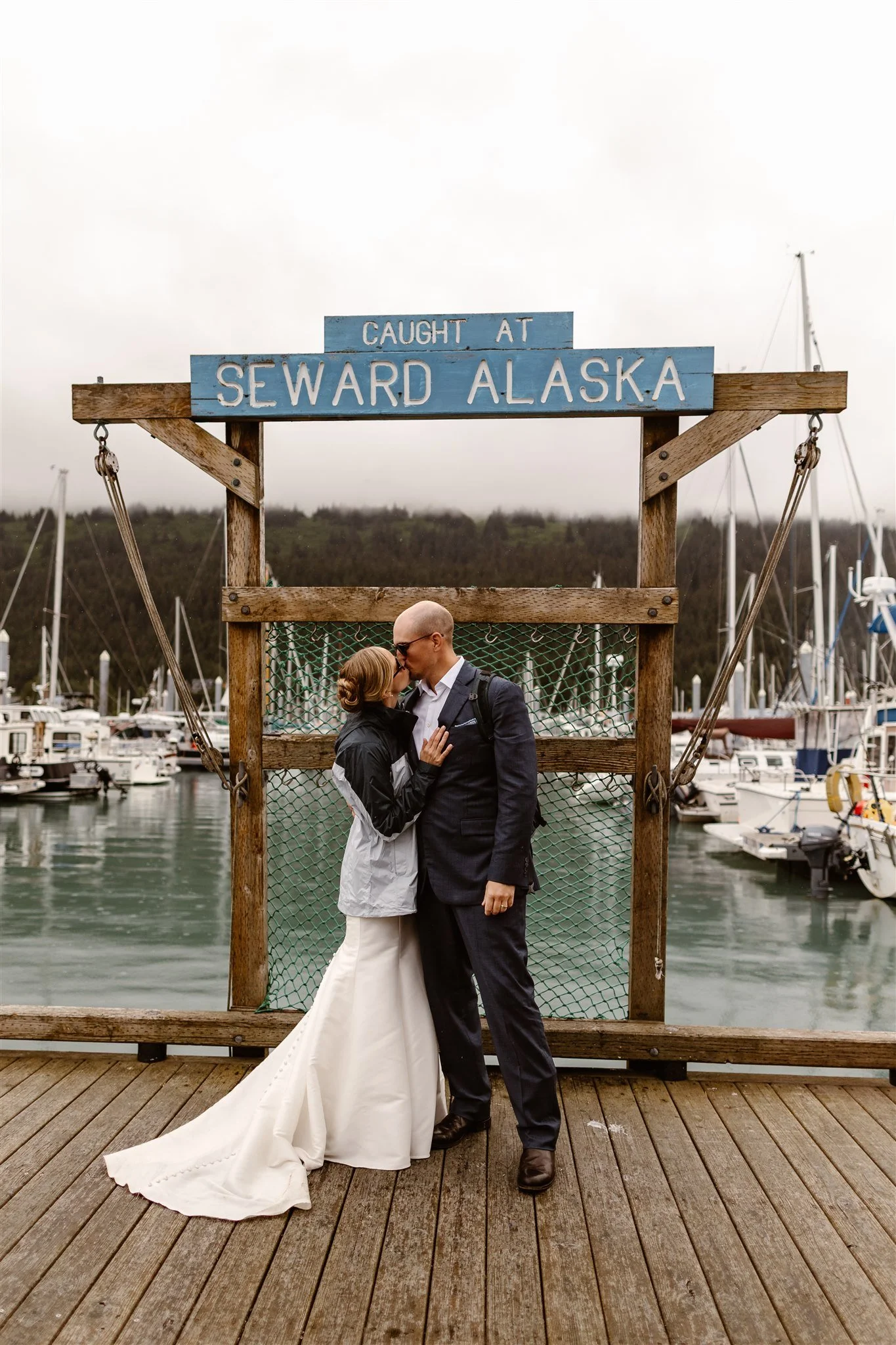 A couple share a kiss in front of the "Caught at Seward Alaska" sign at the harbor as they celebrate their marriage. The bride is wearing a jacket over her wedding gown and the groom is wearing a dark blue suit.