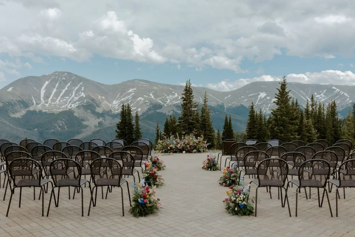 The stone patio of Lunch Rock that has been transformed into a wedding ceremony with an expansive view of the Rocky Mountains. There are rows of metal chairs arranged on the stones and colorful flower arrangements along the aisle and at the altar.