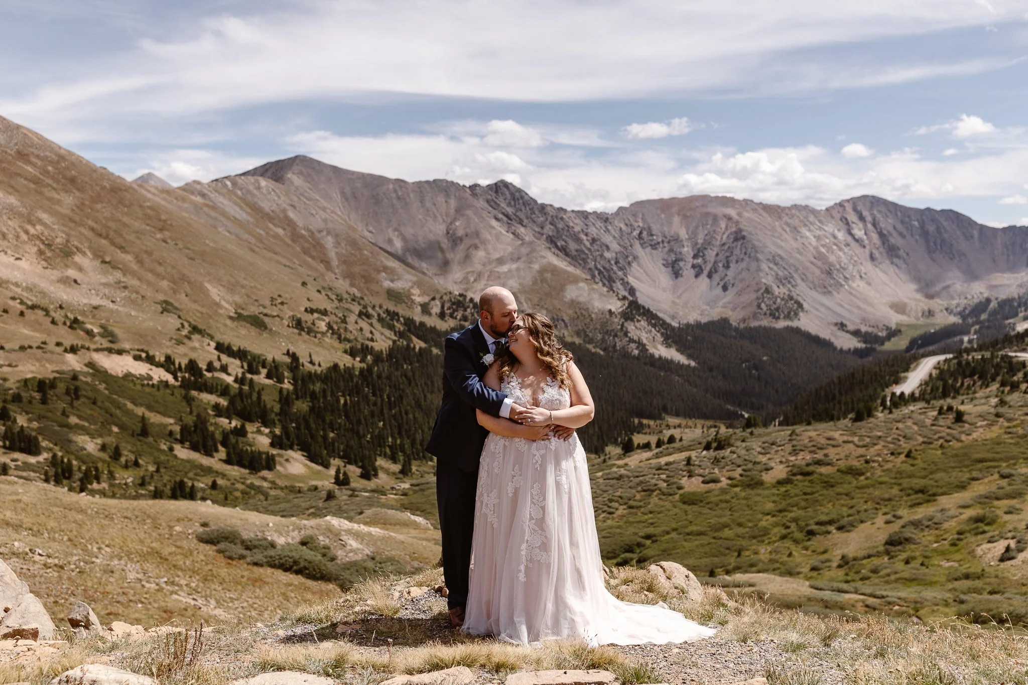During their summer elopement in Breckenridge, CO, the groom holds his bride from behind and kissing her forehead and she smiles back at him with her eyes closed in a very tender moment shared amongst the lush landscape and grey mountains behind them