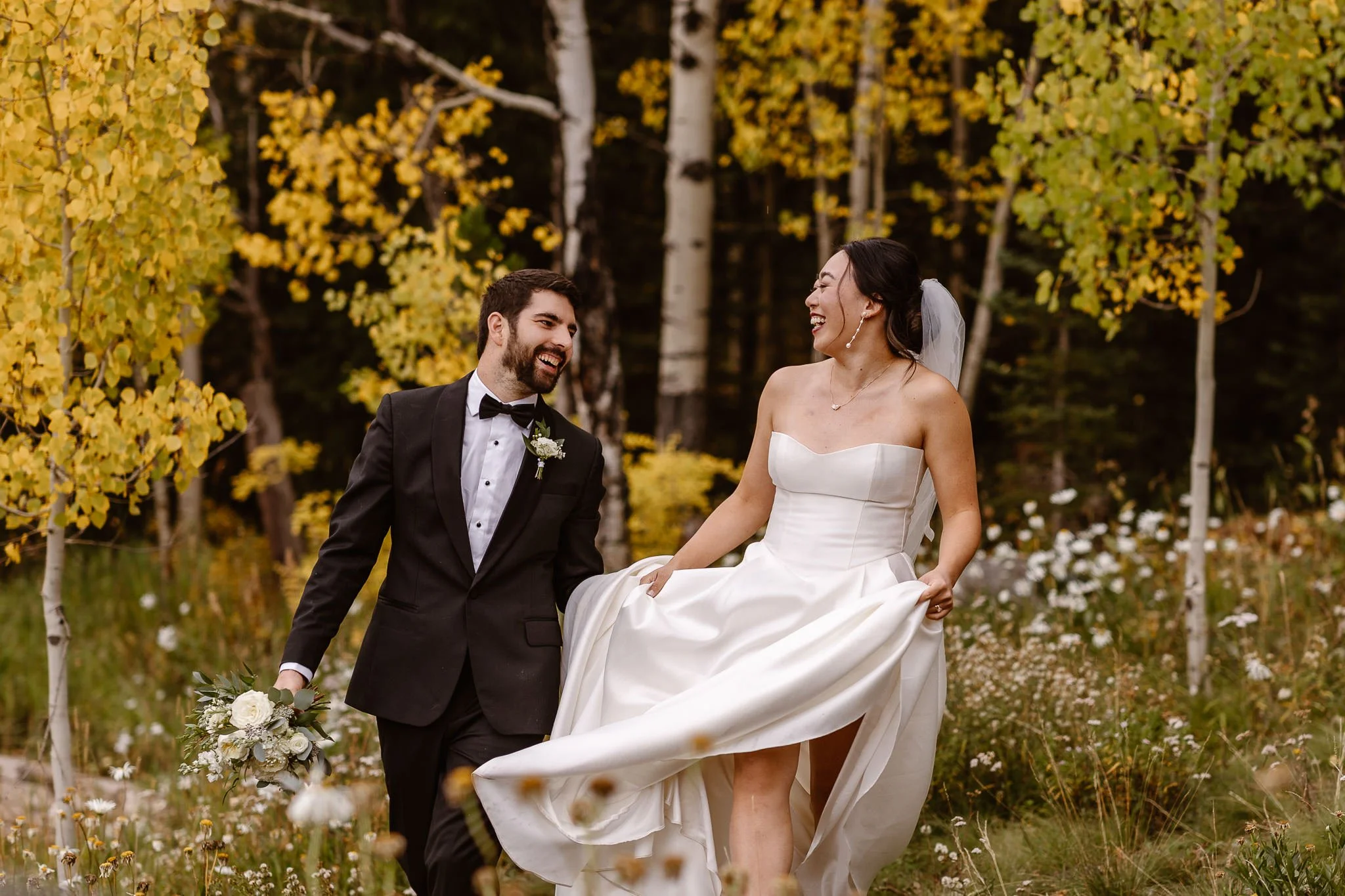 As they run through the field of white flowers in their dress and tux, the newlywed bride and groom smile widely at each other with that "just married" glow. The trees behind them are bursting with yellow as fall settles into the landscape.