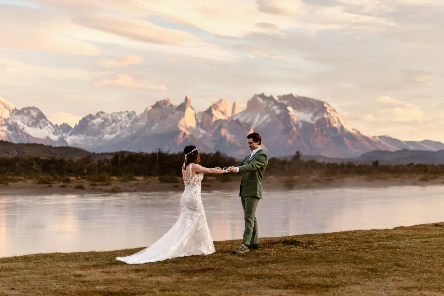 During their sunrise first look, the groom, in a green suit, holds the bride's hands as he stands back and takes her in. She's wearing a lace, open back trumpet wedding gown as the mountains glow in pink morning light behind them.