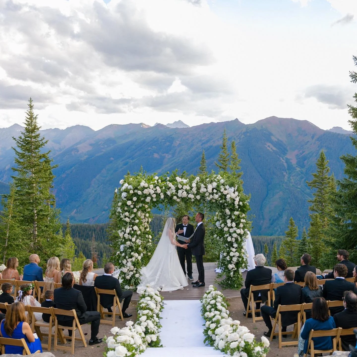A sunset wedding ceremony on the Aspen Wedding Deck , decorated beautifully with greenery and white blooms all around the square arch and on either side of the aisle. The guests are seated in wooden folding chairs and the mountains fade in the light.