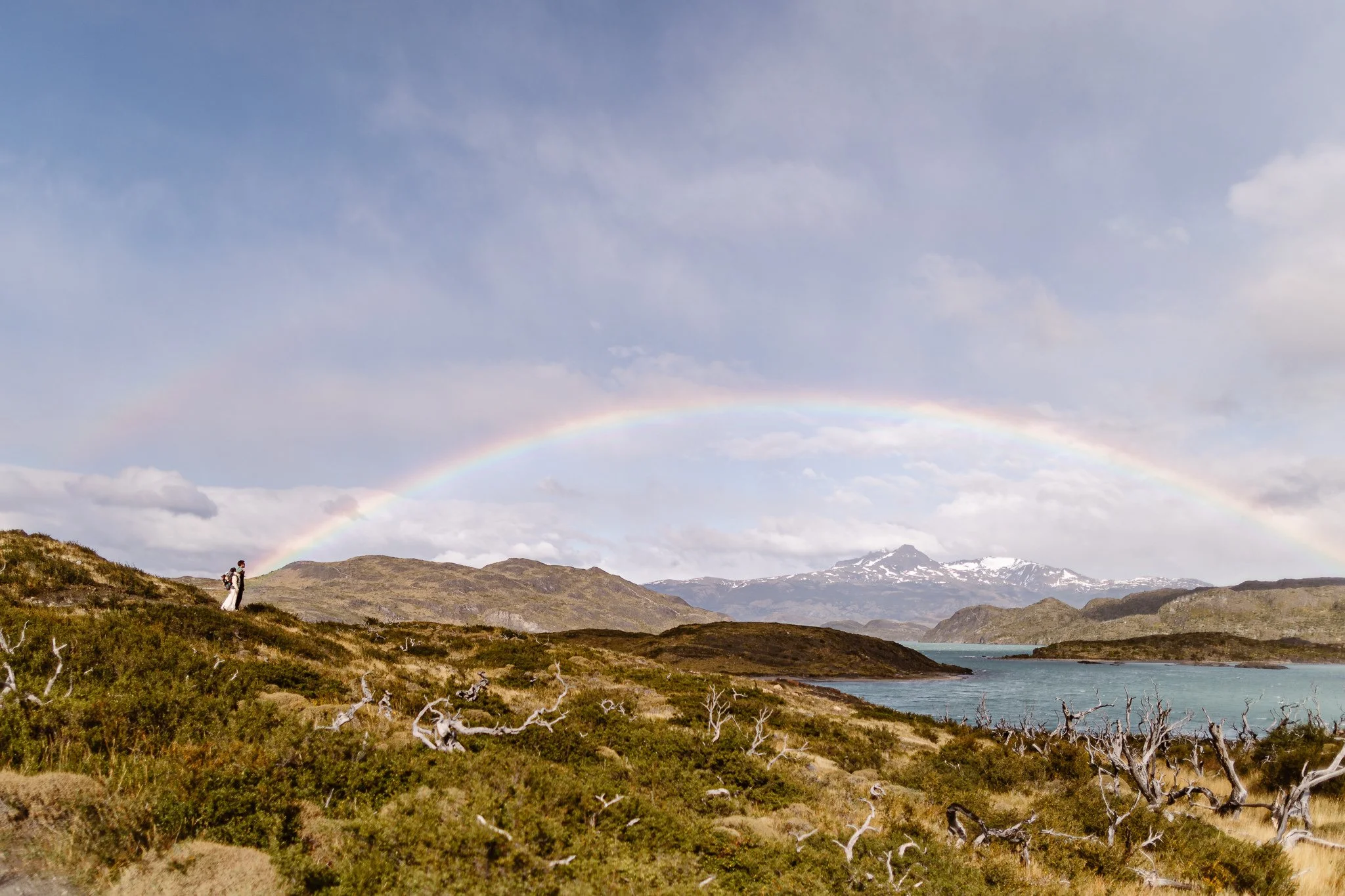 Newlyweds stand at the end of a rainbow near a blue lake in Patagonia with dramatic mountain peaks behind them