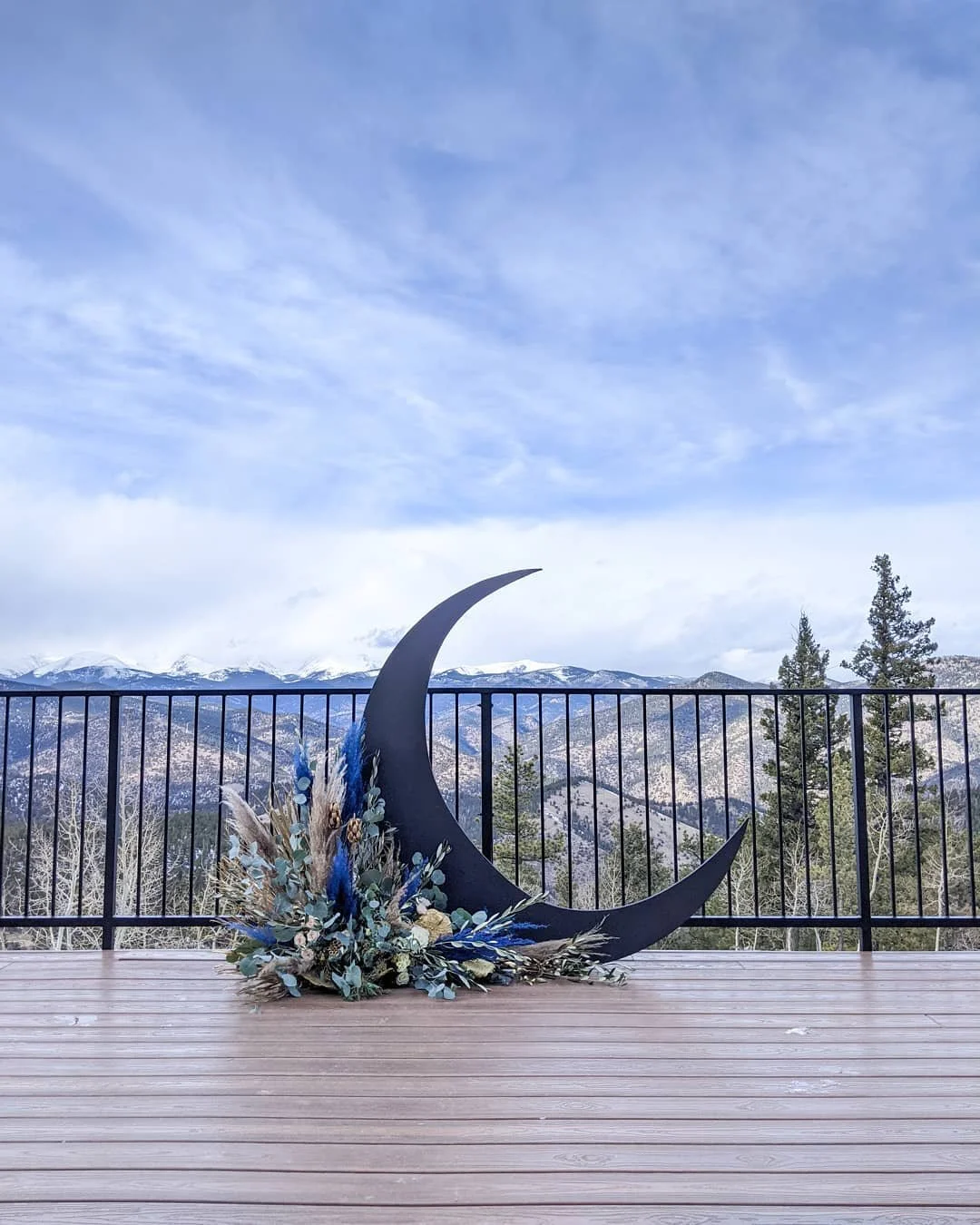 The outdoor wedding deck at North Star Gatherings where you can hold your ceremony on a simple wooden deck with a metal barrier that allows you to see the Rocky Mountains in the background. On the deck sits a black crescent moon with florals decor