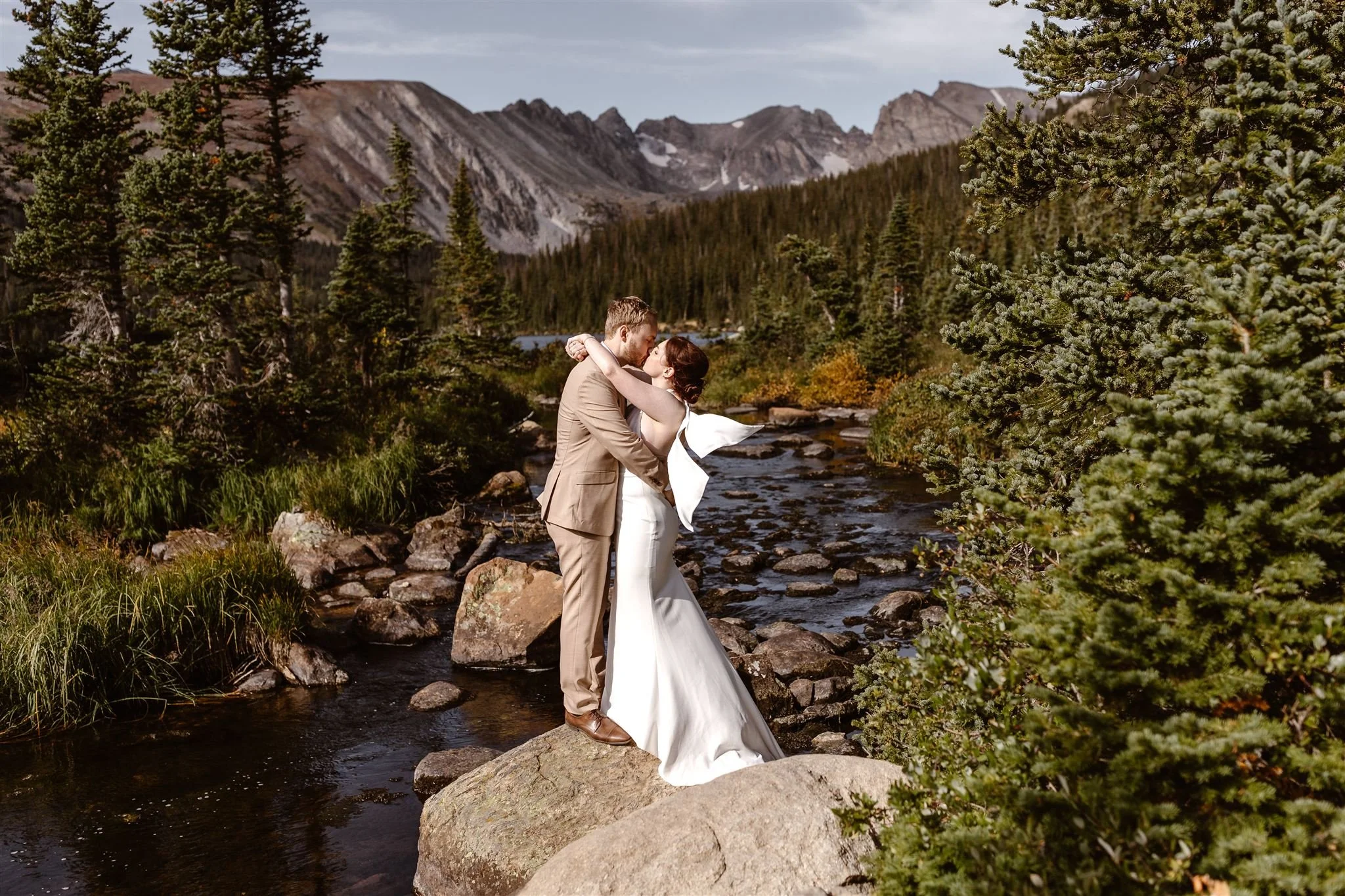 The bride and groom holding each other while embracing in a romantic kiss on top of a rock on the calm creek near Lake Isabelle. The mountains fill the background as the the lush green trees surround them on all sides.