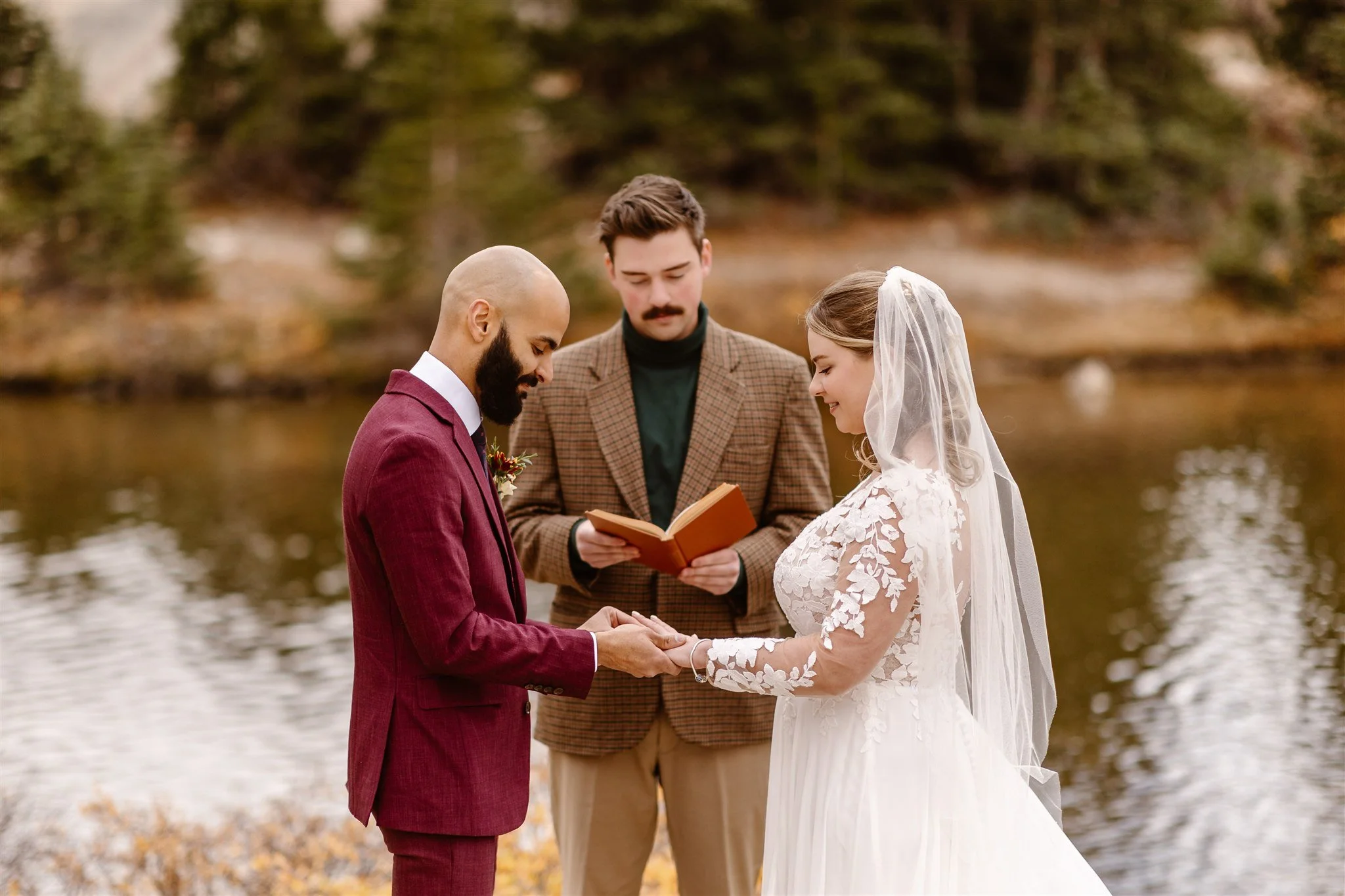 As the groom places the ring on his bride, the two look down as her hand adoringly and their officiant reads from a brown leather book behind them. The lake shimmers in the background and the fall colors are in full effect.