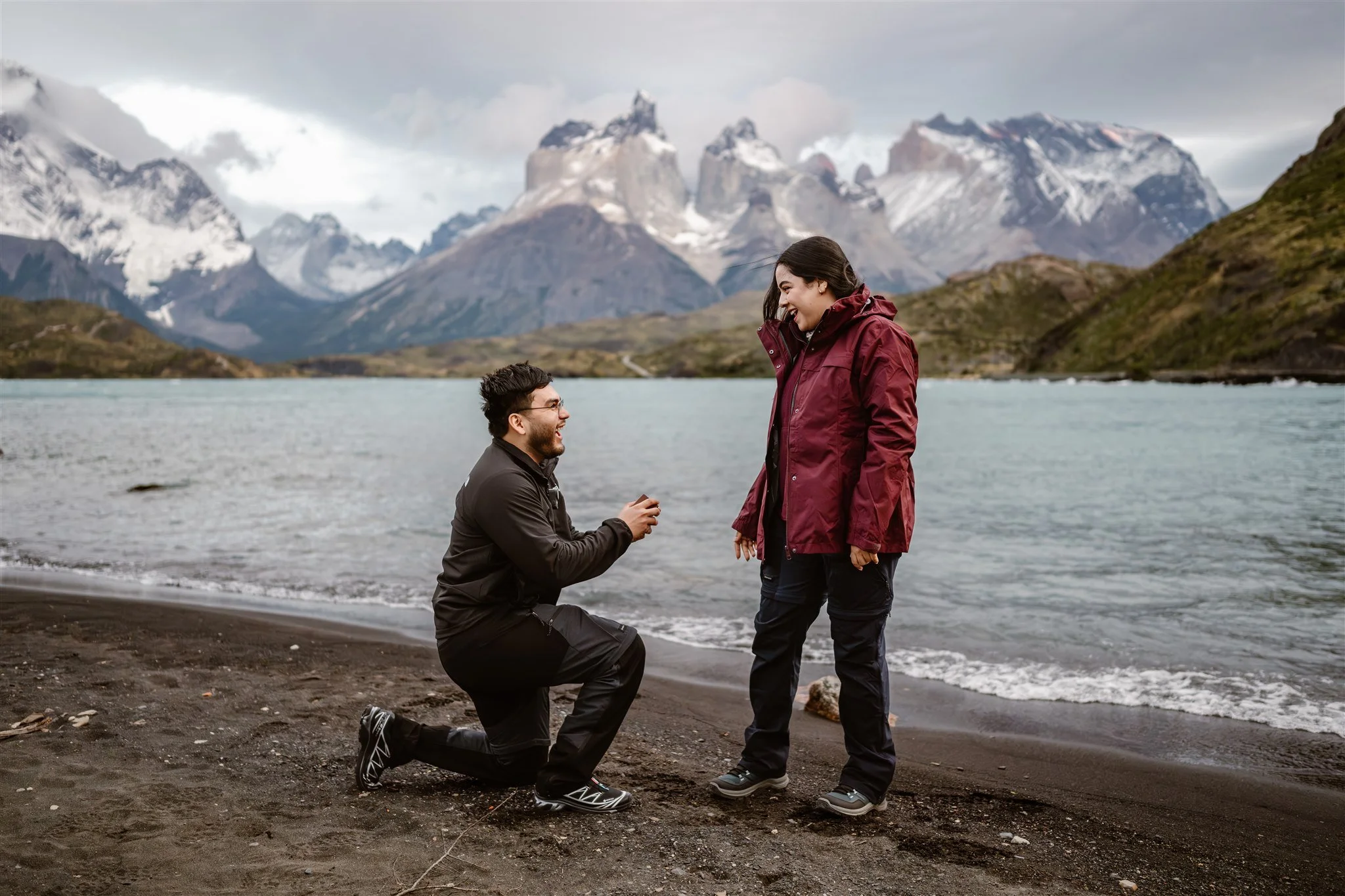 On the coast of the turquoise waters of Torres del Paine in Patagonia, a young man holds out a ring box while kneeling on one knee as he asks his girlfriend to marry him. She looks at him in shock with a smile on her face as the wind blows their hair
