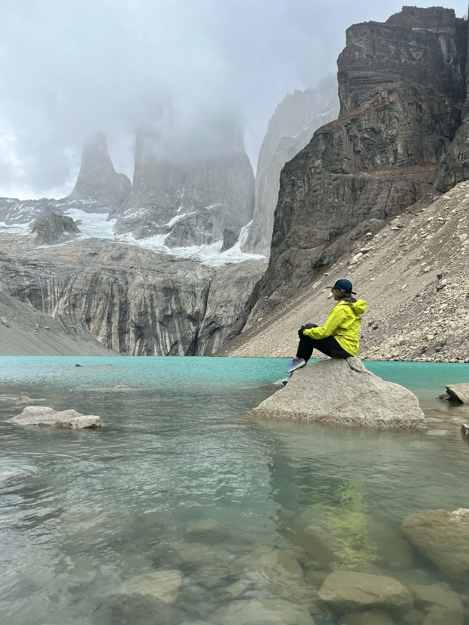 Elopement photographer, Andrea Enger, wears a bright yellow/green jacket while sitting atop a rock in the middle of a turquoise lake while dramatic rock formations jut out of the water