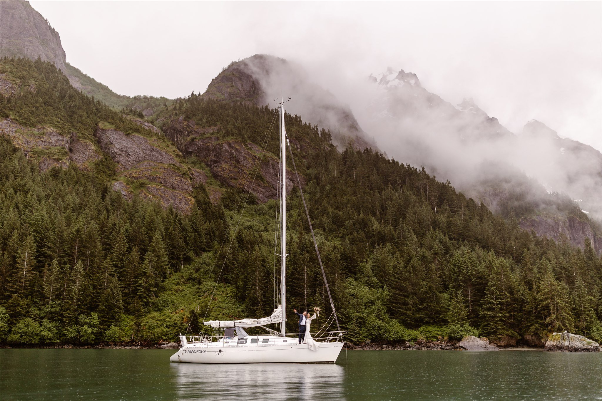 As the mist covers the top of the towering, tree-covered mountains behind them, a newly-married couple pose on the edge of the sailboat during their epic elopement.