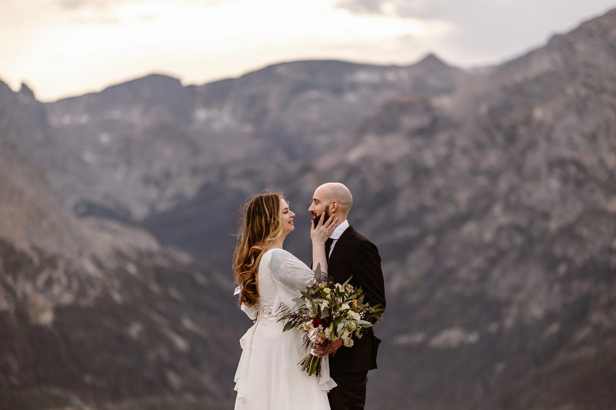During a joyful moment, the bride uses her right hand to gently hold her partner's beard and they smile at each other. The mountains and evening sky fill the background as the colors turn cool and even more romantic.