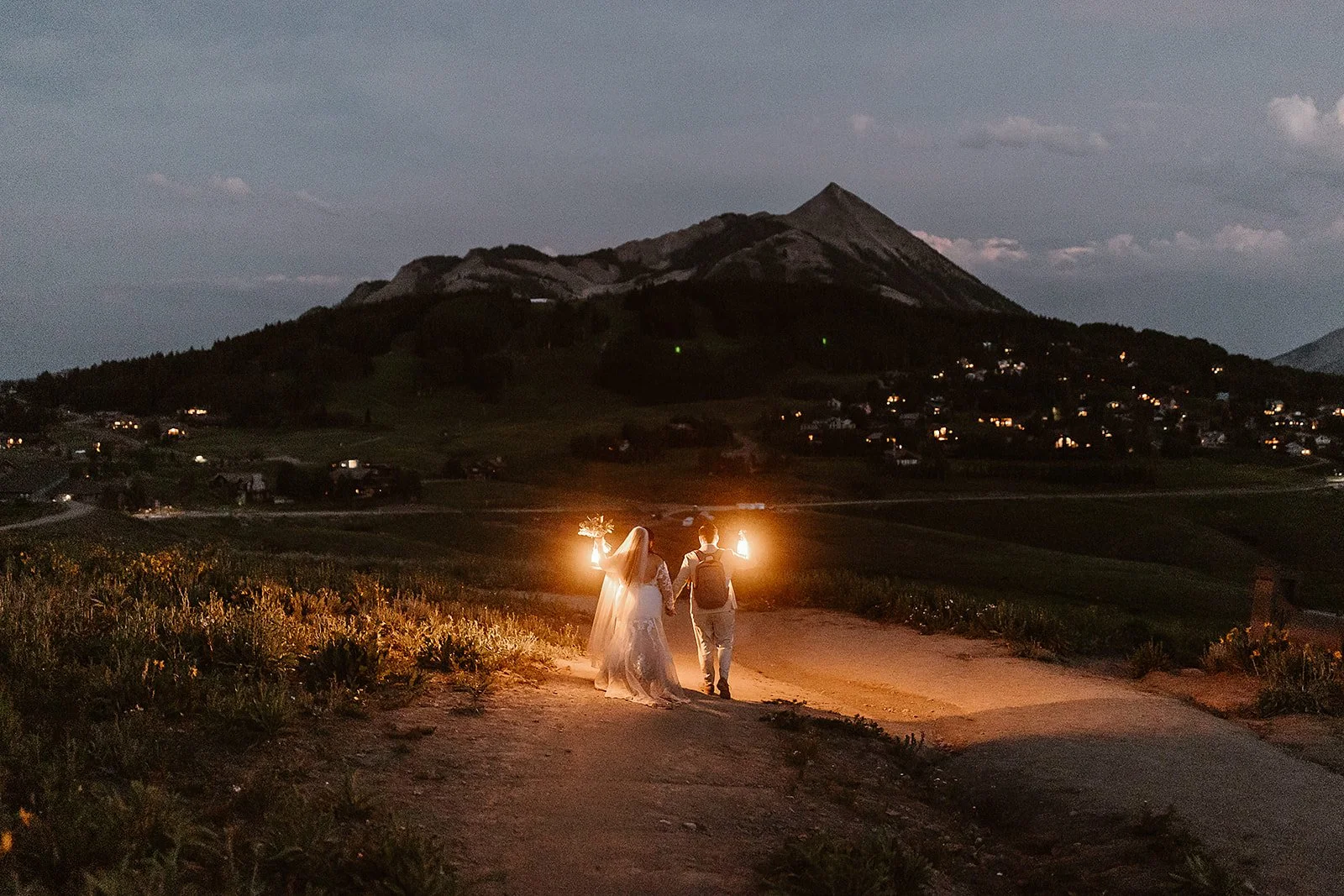 Newlyweds walking back to town at the end of their gorgeous summer elopement picnic in Crested Butte during wildflower season. Mount Crested Butte fills the background, as their lanterns light the way before them.