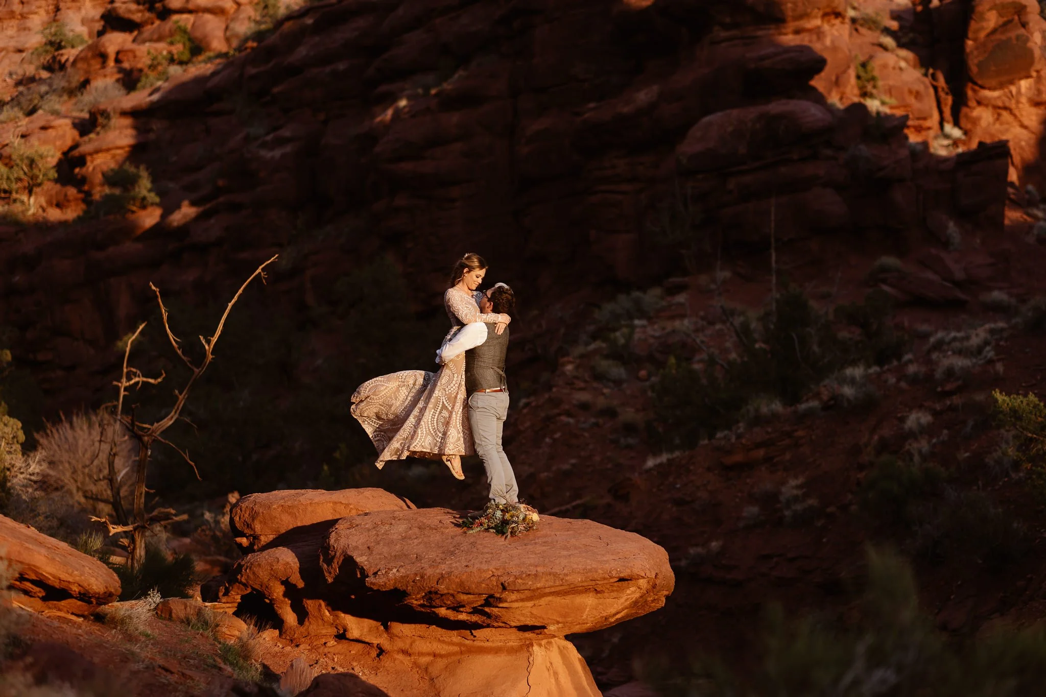 Freshly married, this bride and groom share a beautiful moment as the groom lifts his wife up into the air, her dress flowing behind her in the wind, as they stand the edge of red rocks, while also surrounded by them during an orange-filled sunset.