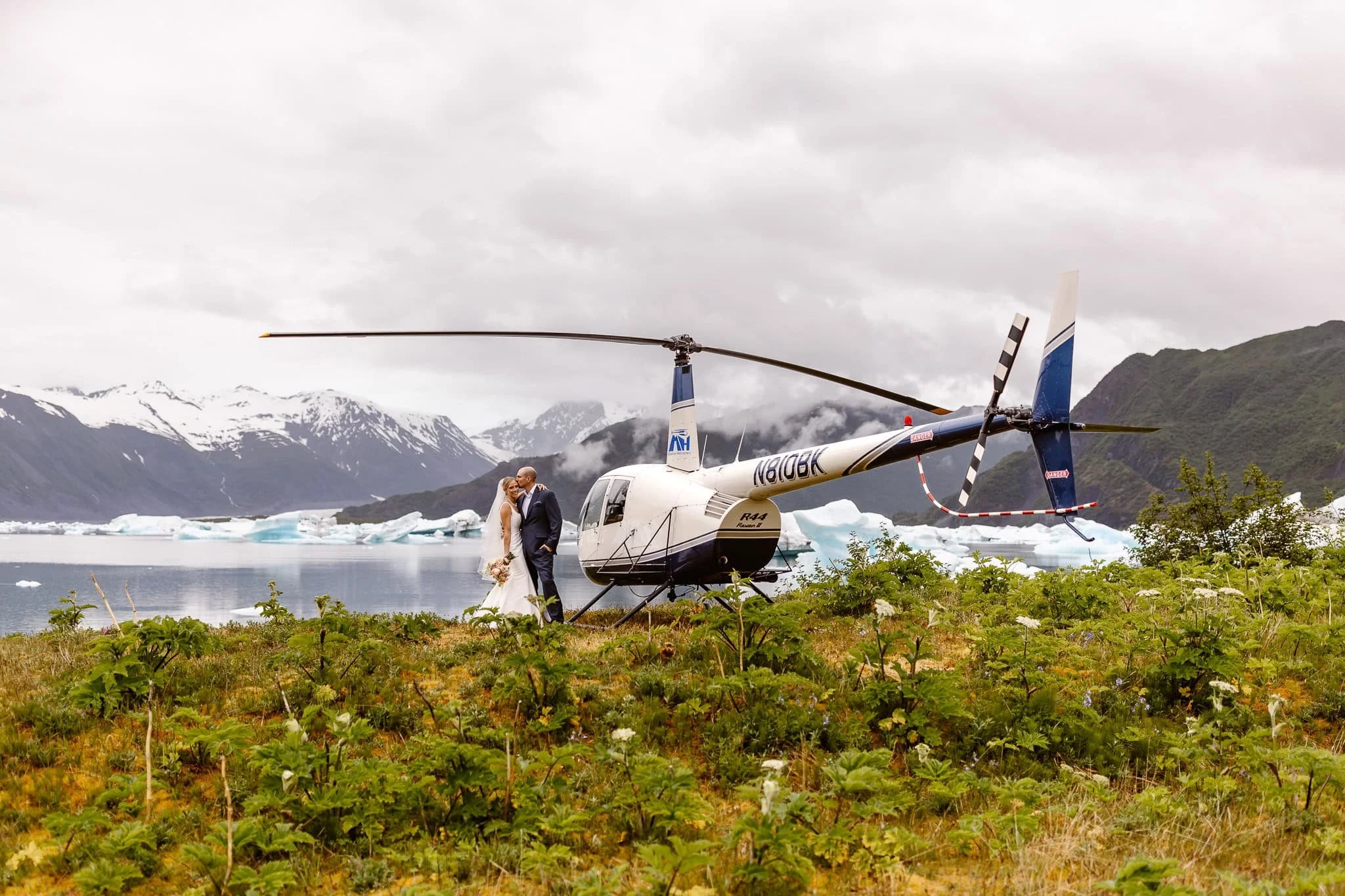 Standing next to the helicopter that flew them to this glacier, the groom kisses his bride’s forehead. The landscape consists of light blue icebergs floating in the water, lush foothills, and snow-capped mountains.