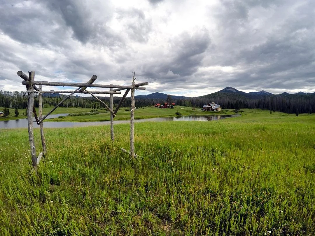 The wooden arch for weddings is situated in the grass near the lake at Midnight Ranch, with the main buildings off in the distance on the other side of the lake, with the mountains in the background.
